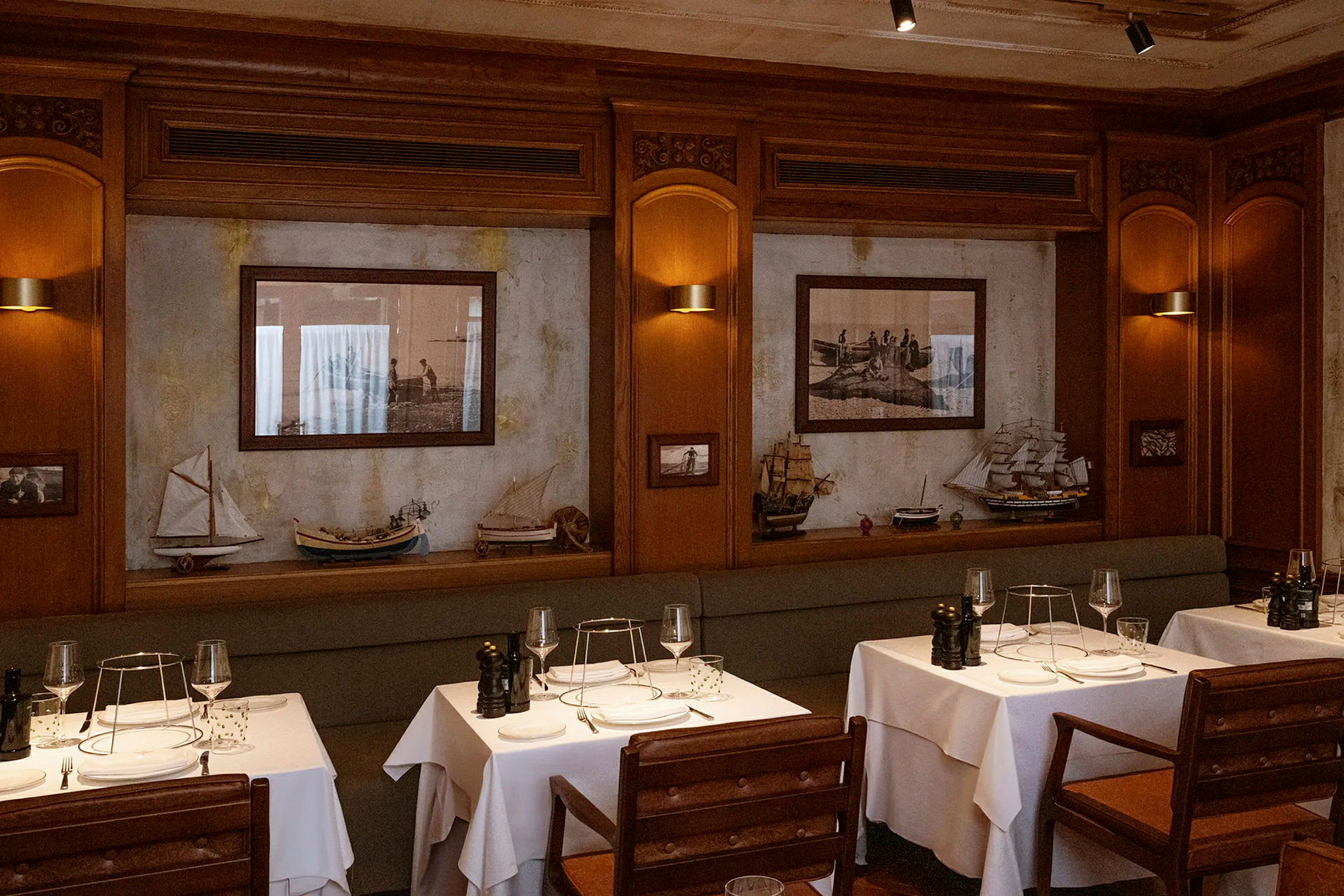 En elegant restauranginteriör med bord dukade med vita dukar och trämöbler. Väggarna pryds av tavlor och modellbåtar. (AI-genererad text)