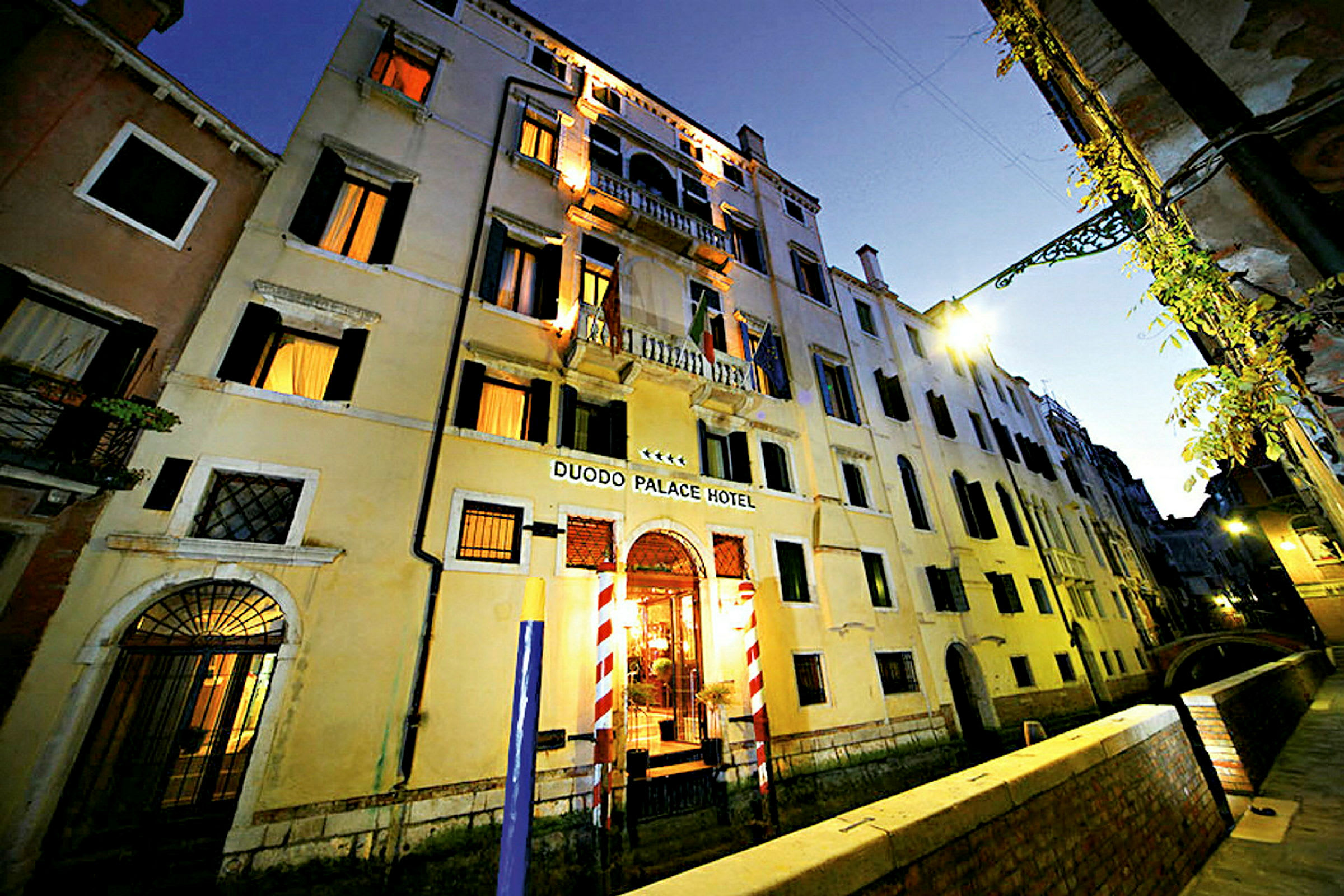 Flerbostadshus i tre våningar med skylt Duodo Palace Hotel, omgivna av en kanal och en gångväg. (AI-genererad text)