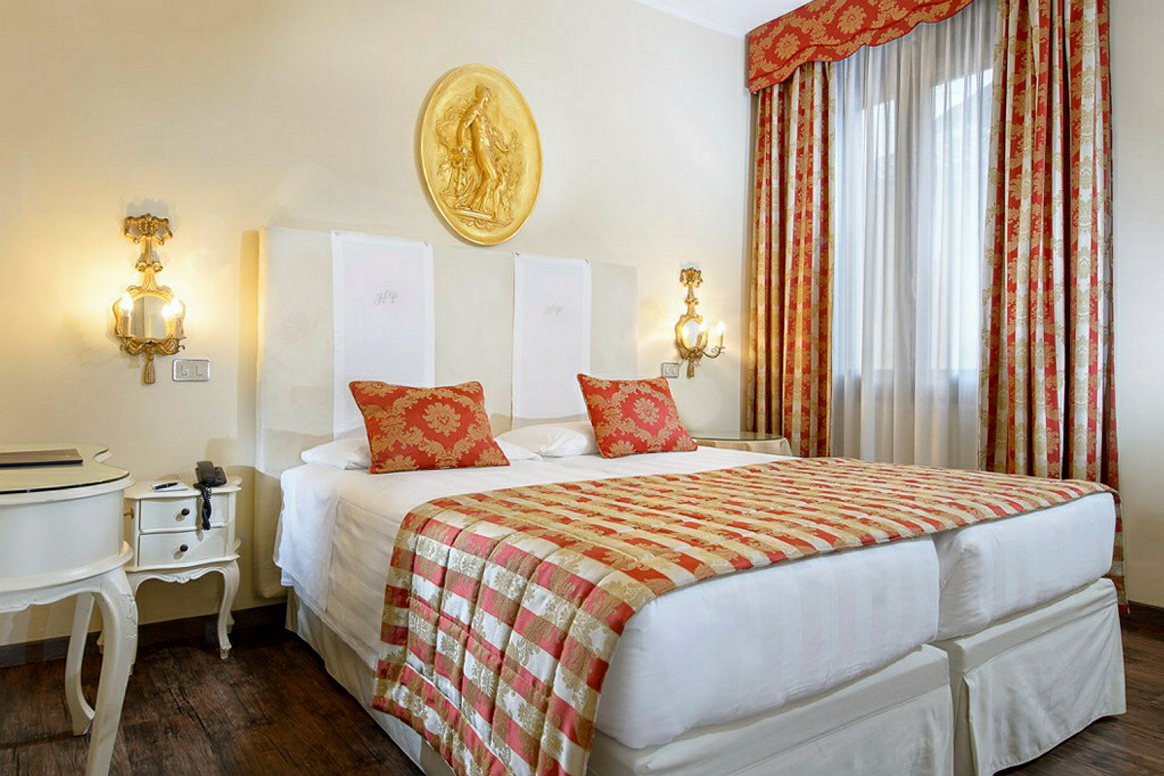 Ett hotellrum med en stor säng dekorerad med röda och gyllene textilier, vita möbler och dekorativ belysning. (AI-genererad text)