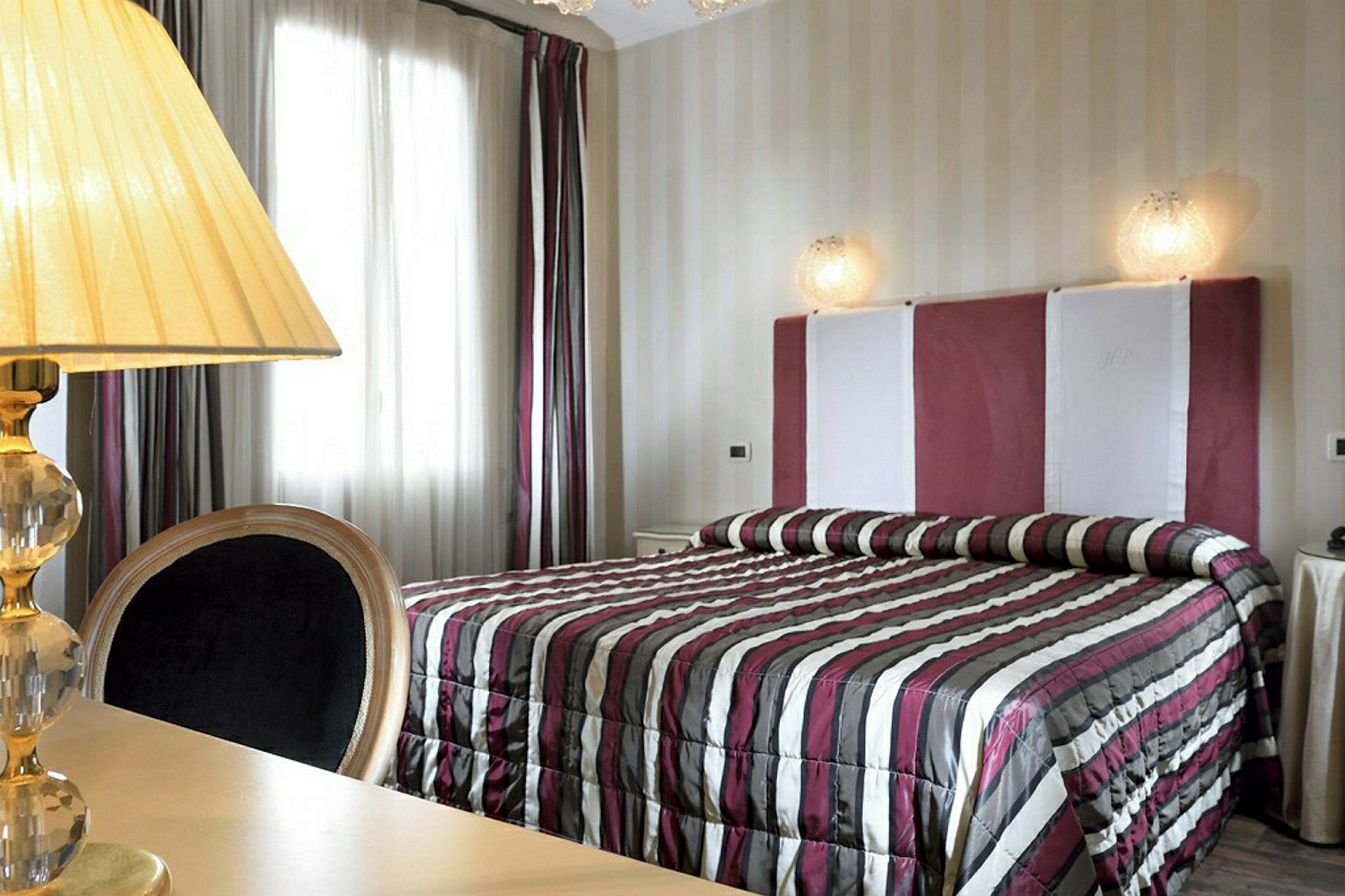 Ett sovrum med en säng som har en randig överkast och en stoppad sänggavel. Till vänster finns en lampa på ett bord. (AI-genererad text)