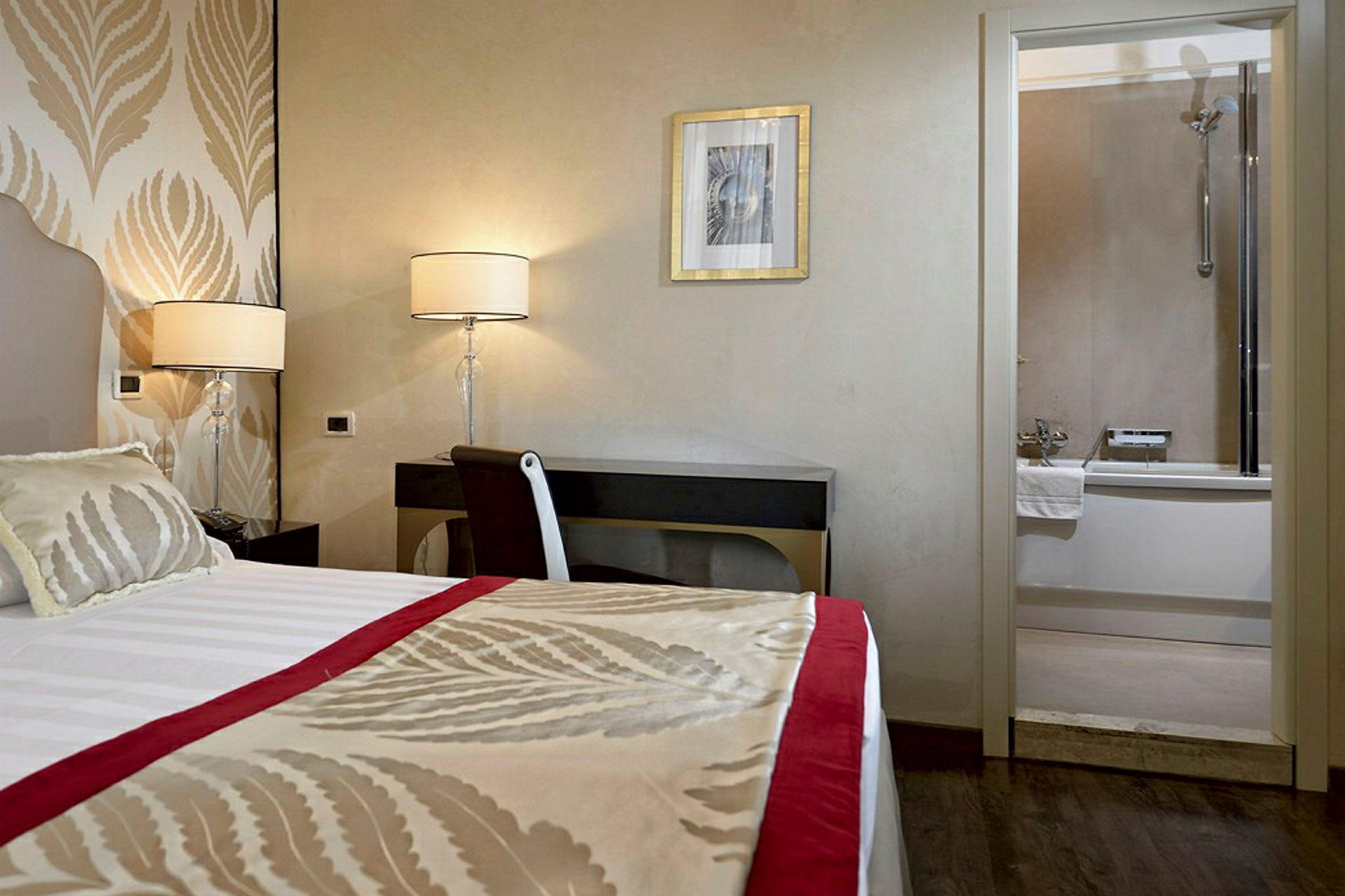 Hotellrum med en säng, skrivbord och lampor, samt ett badrum med badkar i bakgrunden. (AI-genererad text)