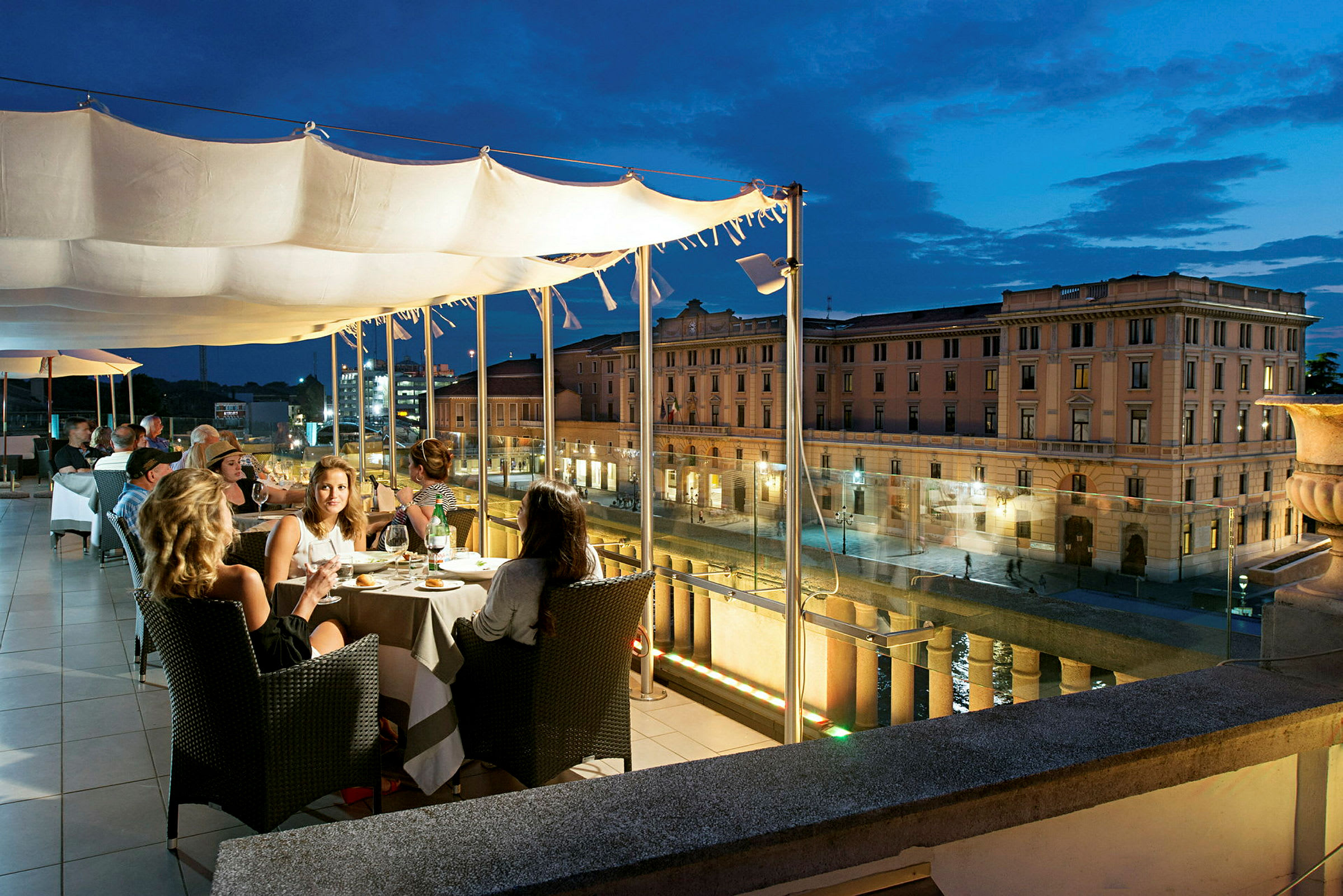 En restaurangterrass på kvällen med utsikt över en stadsgata och en klassisk byggnad i bakgrunden. (AI-genererad text)