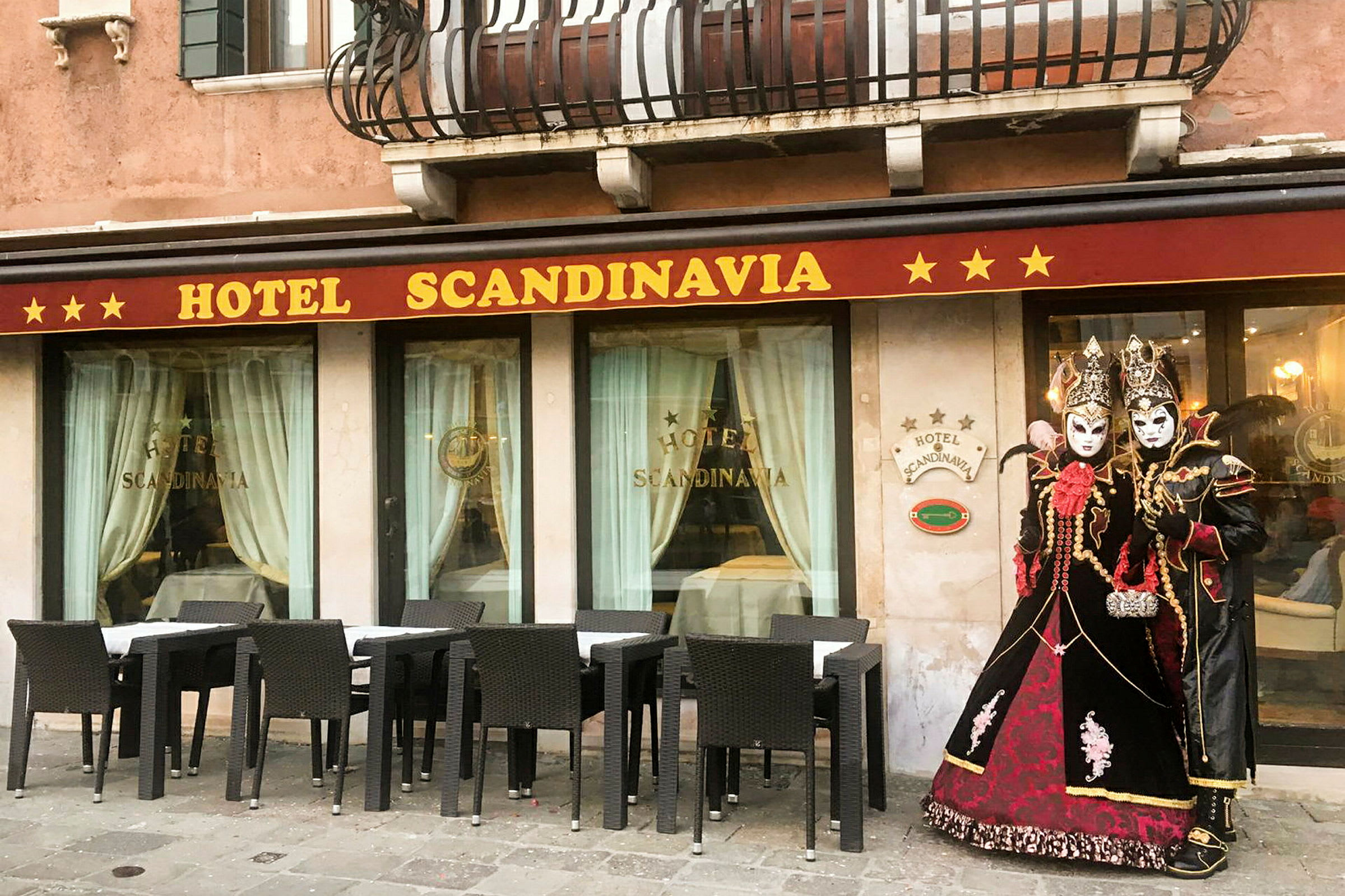 Hotellfasad med namnet 'Hotel Scandinavia', möblerade sittplatser utomhus och personer klädda i traditionella maskeradkläder. (AI-genererad text)
