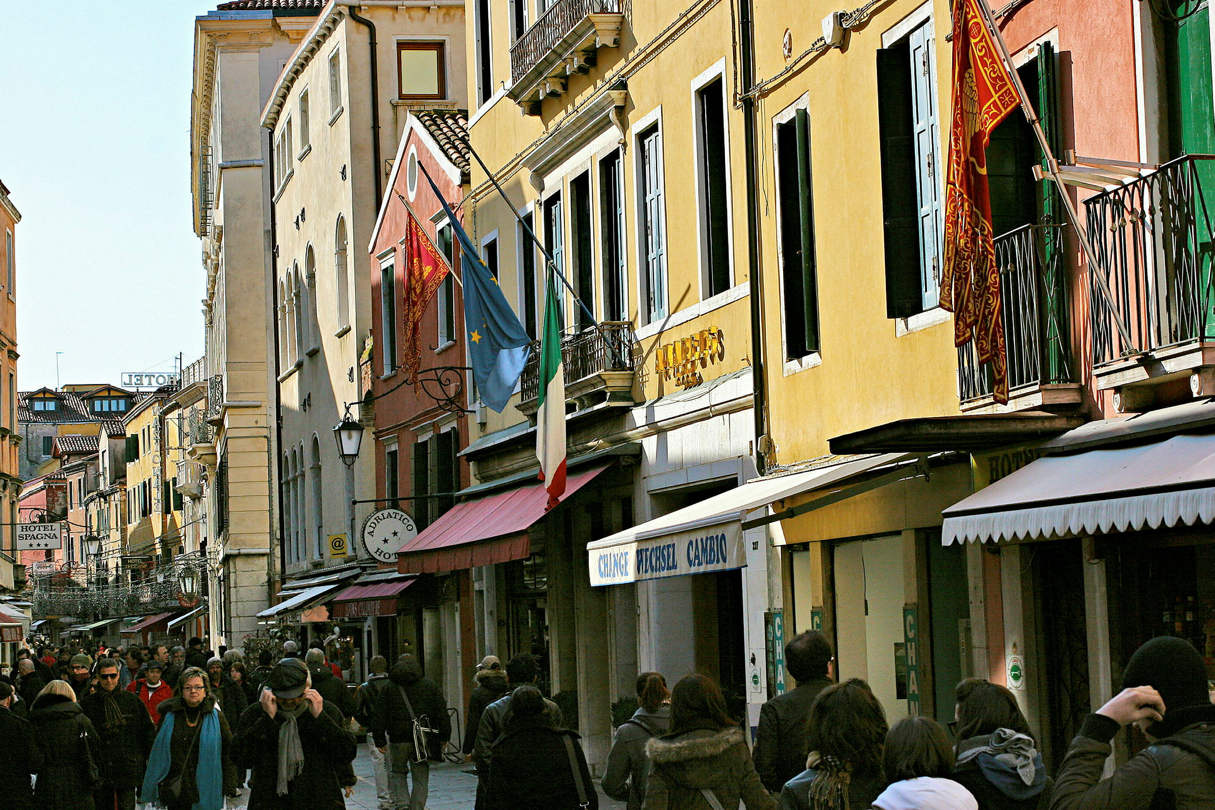En livlig gata kantad av färgglada byggnader med italienska och europeiska flaggor, omgiven av människor som promenerar. (AI-genererad text)