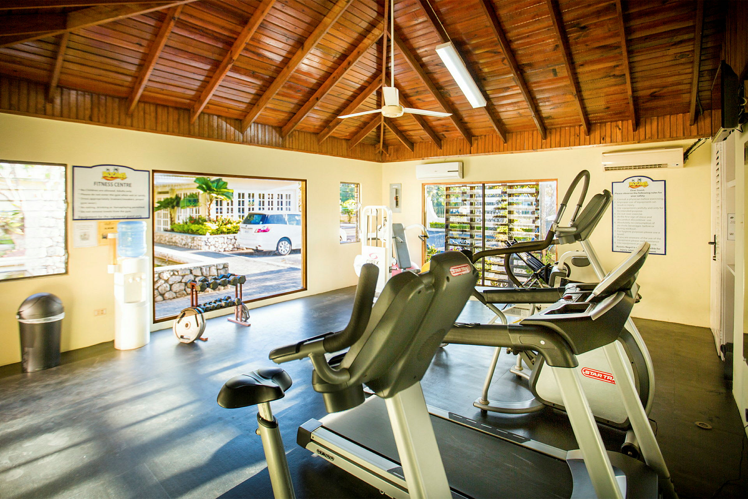 Interiör av ett gym med träningsmaskiner och vikter, träpaneler på taket och stora fönster. (AI-genererad text)