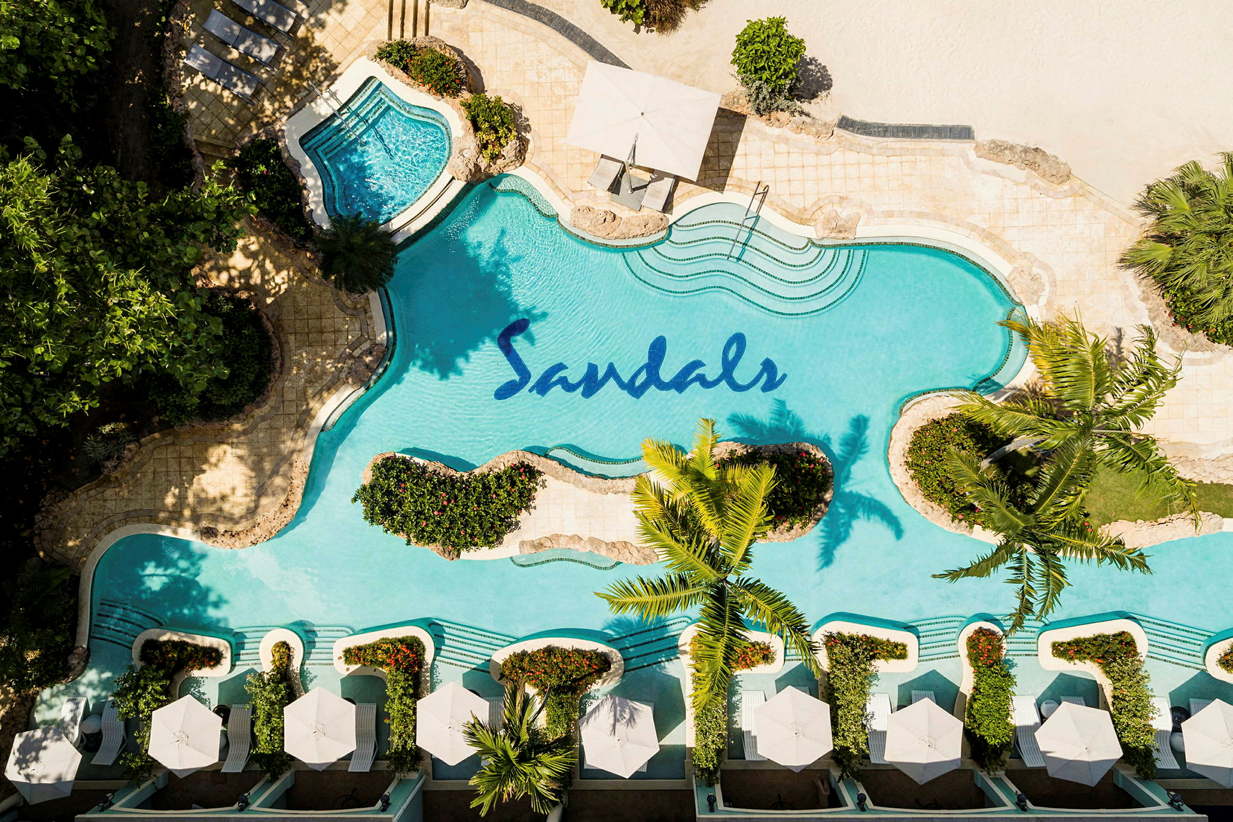 En fågelperspektiv av en pool med Sandals-logo, omgiven av växter och solstolar med parasoller. (AI-genererad text)