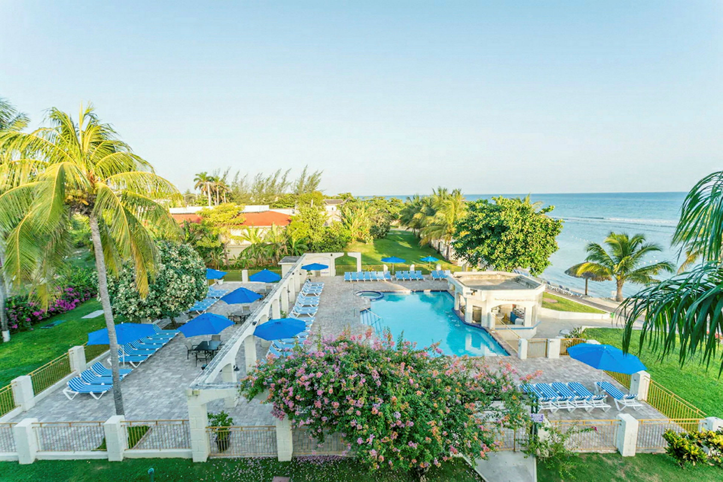 Bild av ett hotellområde vid havet med pool, solstolar och parasoller omgivna av grönska och palmer. (AI-genererad text)