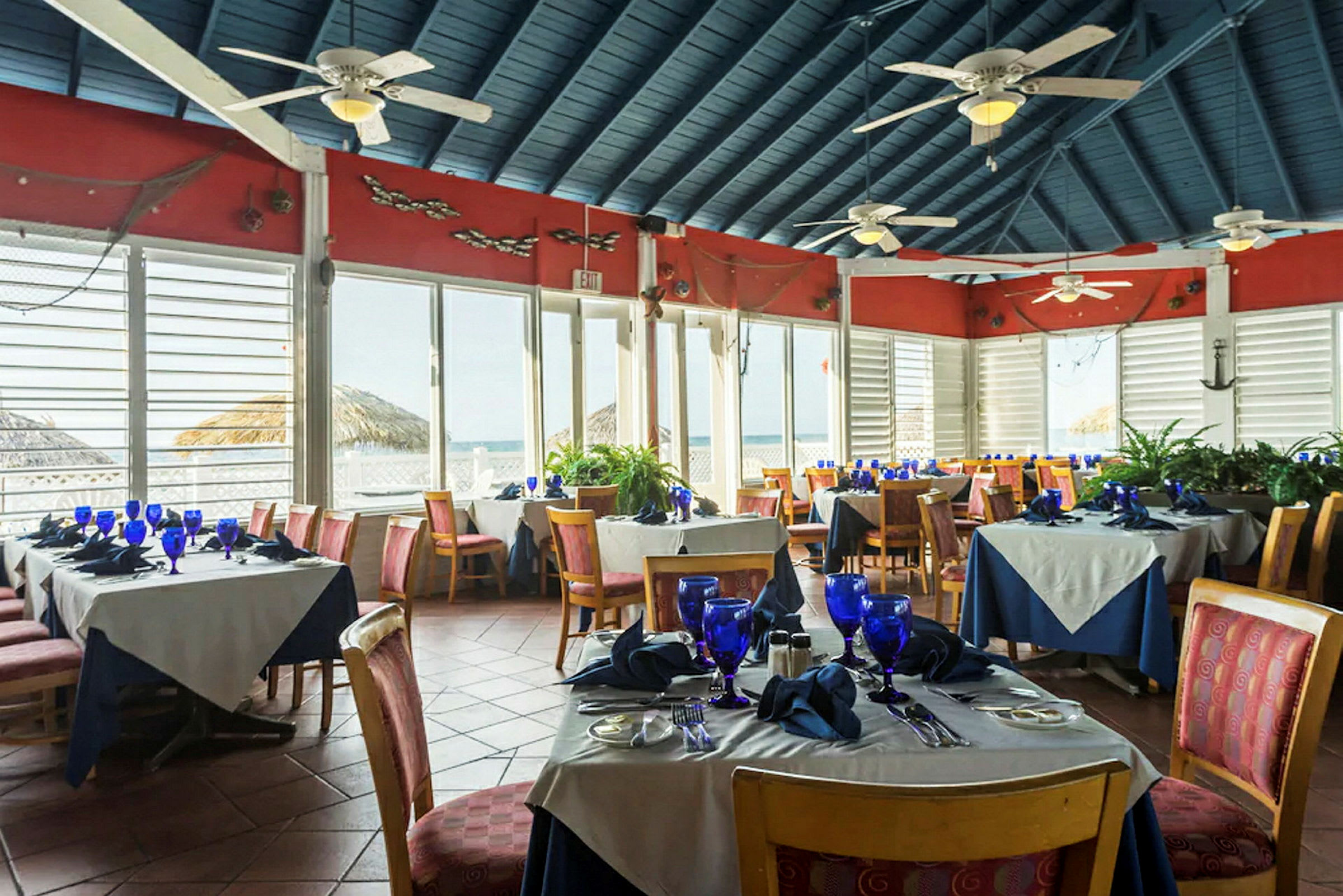 En restaurang med dukade bord, blåa glas och servetter, belägen i ett utrymme med stora fönster och utsikt över havet. (AI-genererad text)