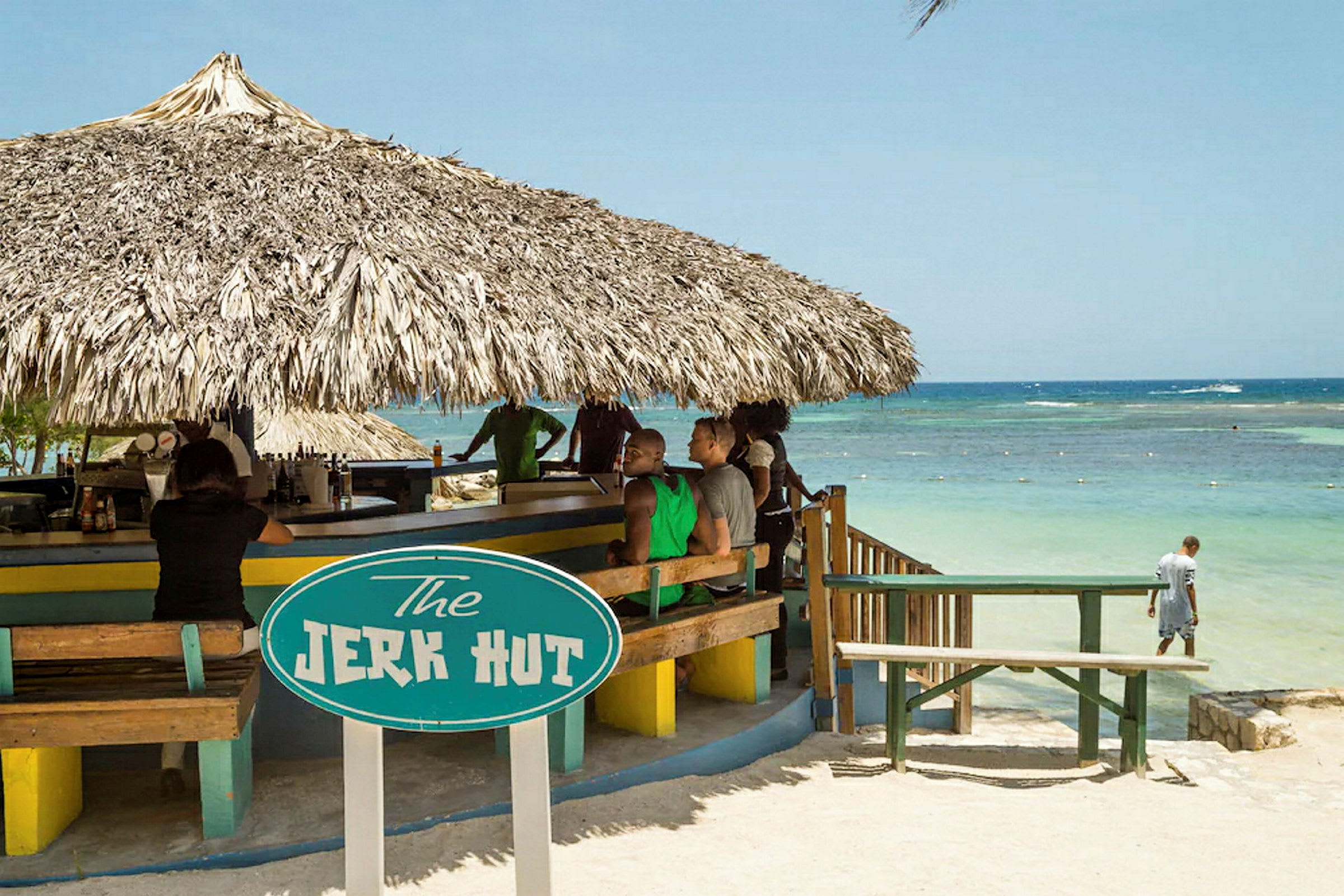 En strandbar med halmtak och en skylt som lyder "The Jerk Hut". Utsikt över havet kan ses i bakgrunden. (AI-genererad text)