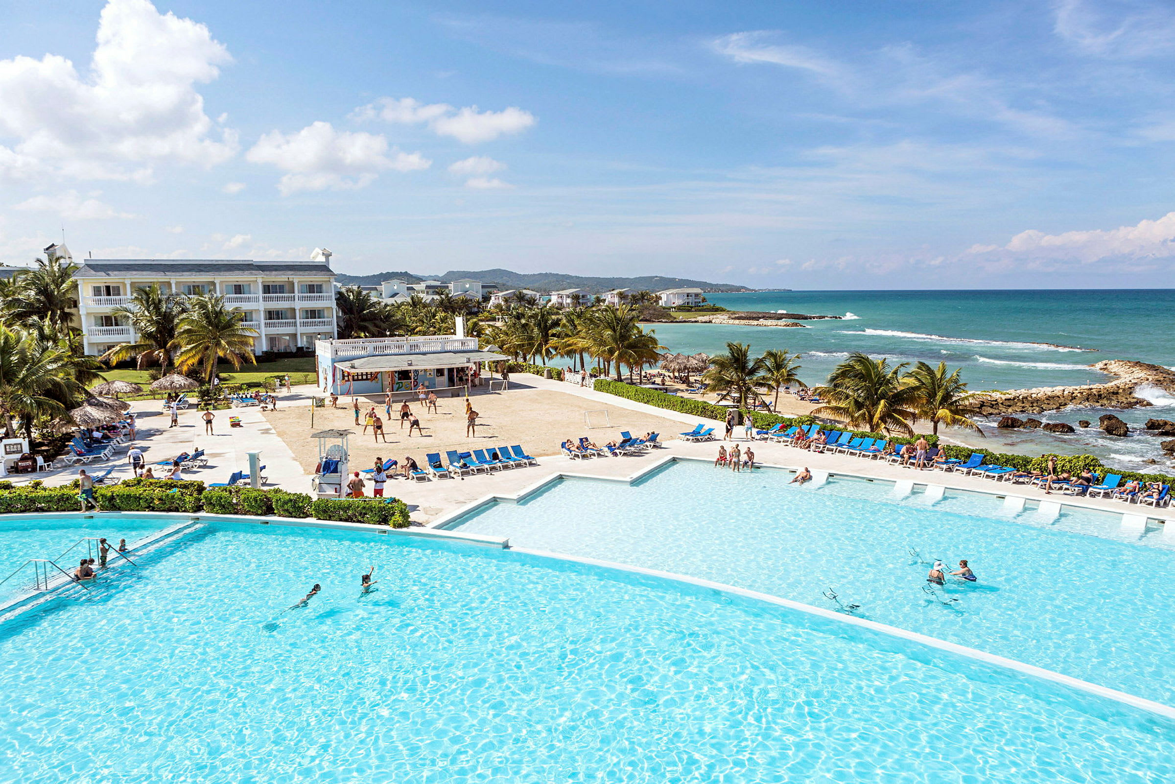 Bild av ett semesterkomplex med en stor pool, solstolar, palmer och en sandstrand vid havet. (AI-genererad text)