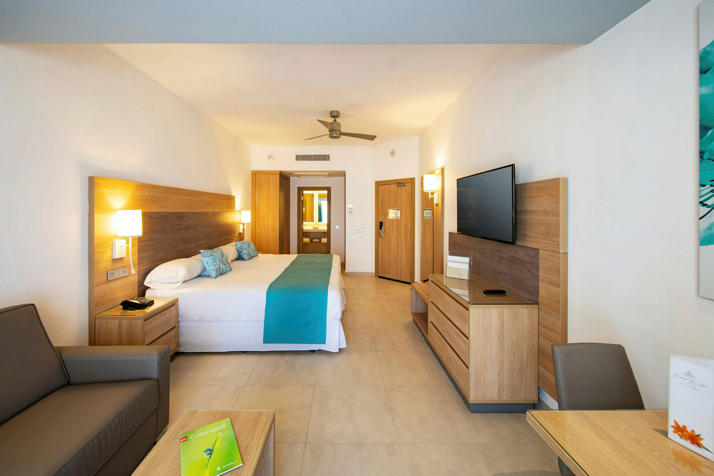 Bild av ett hotellrum med en stor säng, TV, bord och stolar. Ljust inredd och modern. (AI-genererad text)
