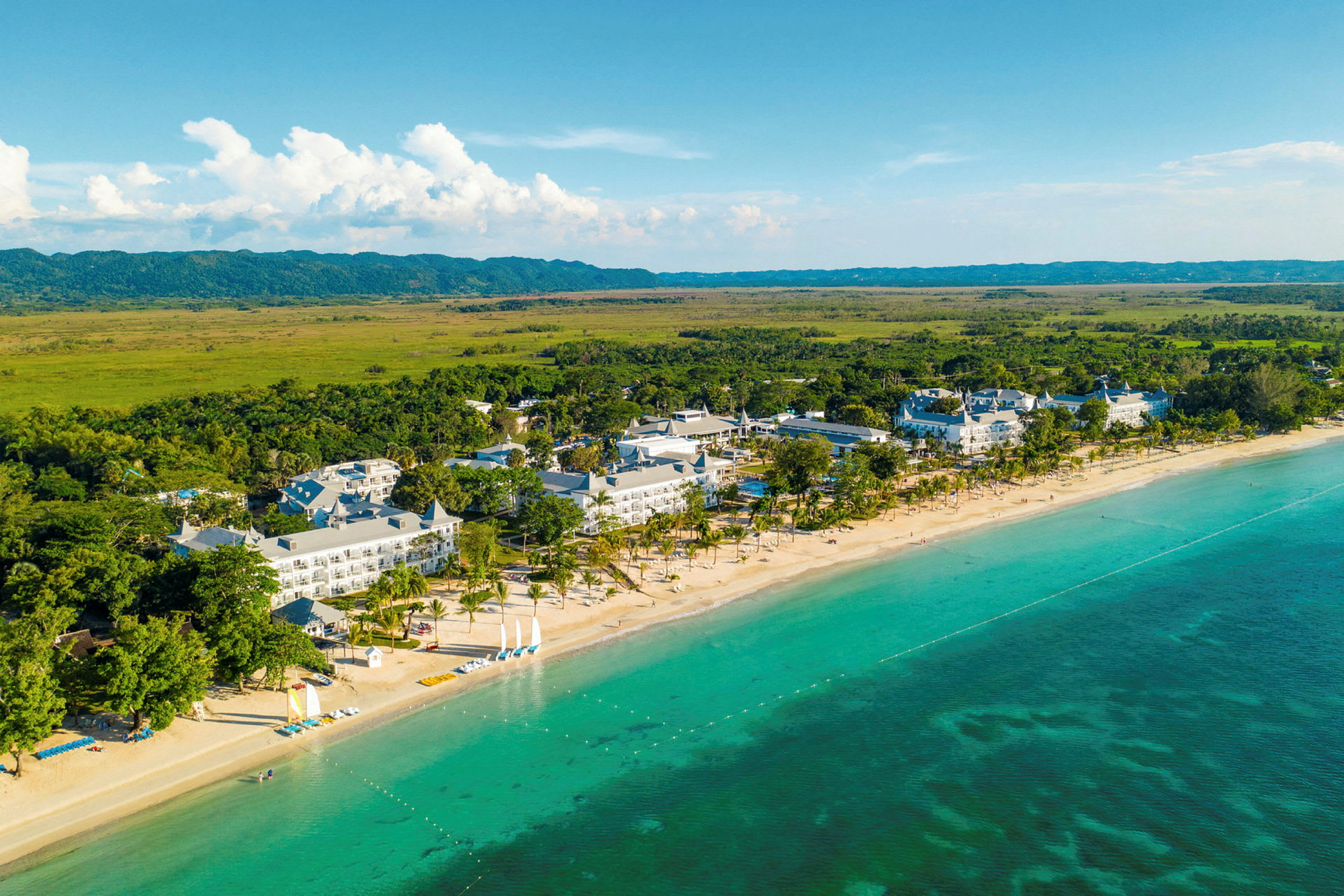 Flygbild av ett kustnära hotellområde med sandstrand, klart turkost vatten och omgivande grönska. (AI-genererad text)