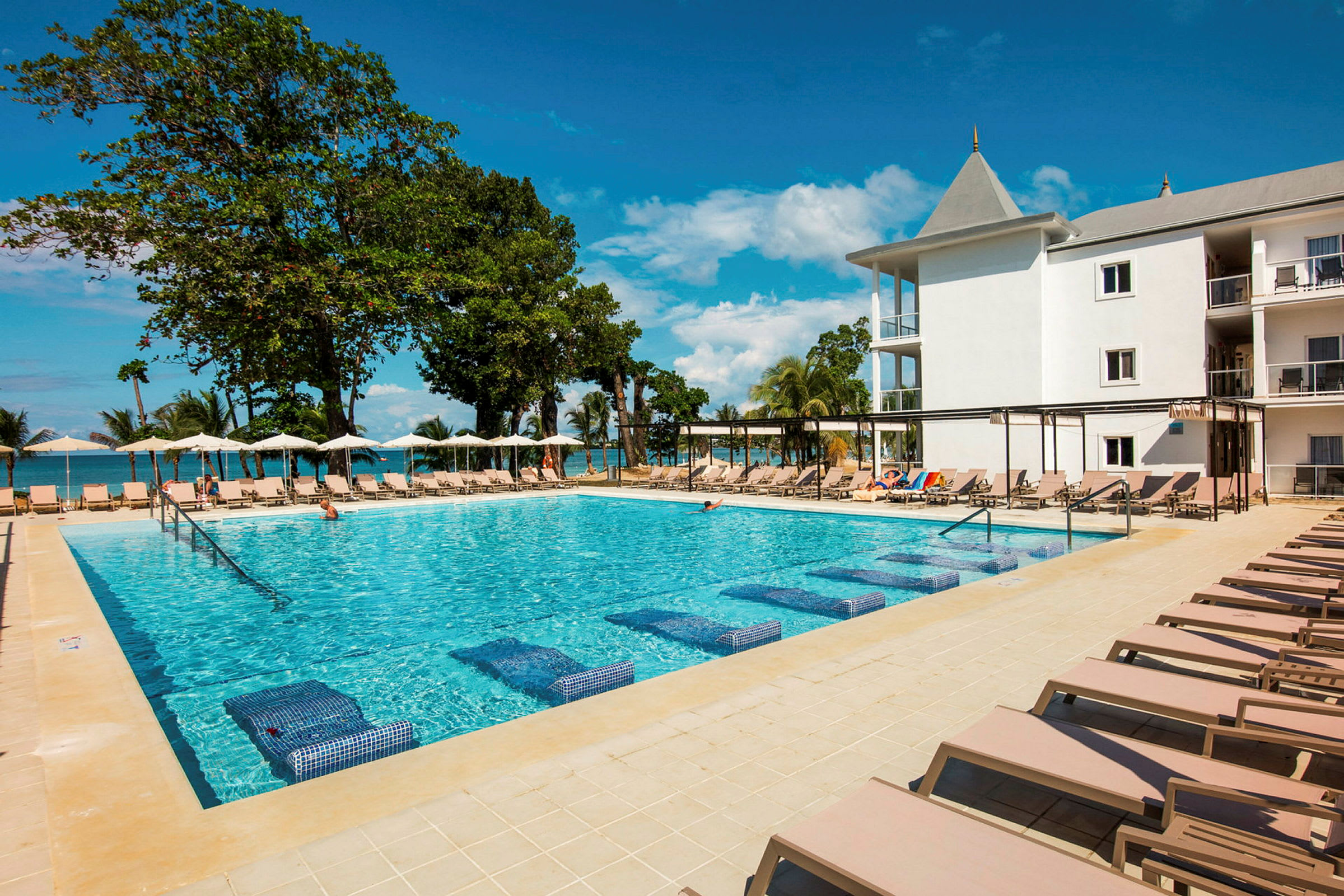 En kristallklar pool omgiven av solstolar med ett hotell och träd i bakgrunden. (AI-genererad text)