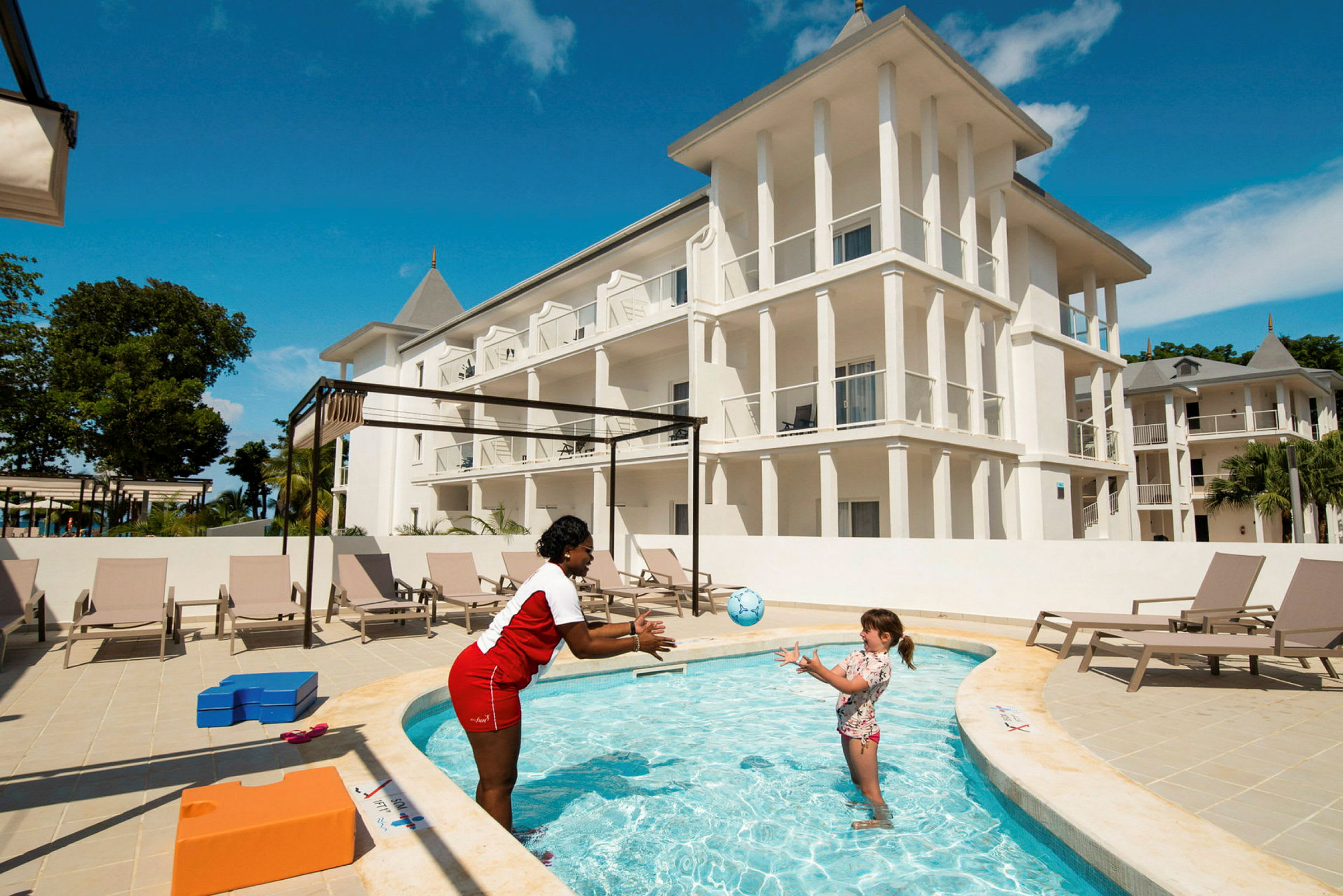 Ett hotell med vit fasad och flera våningar, framför en pool där en person kastar en boll till ett barn. Solstolar står uppställda nära poolen. (AI-genererad text)
