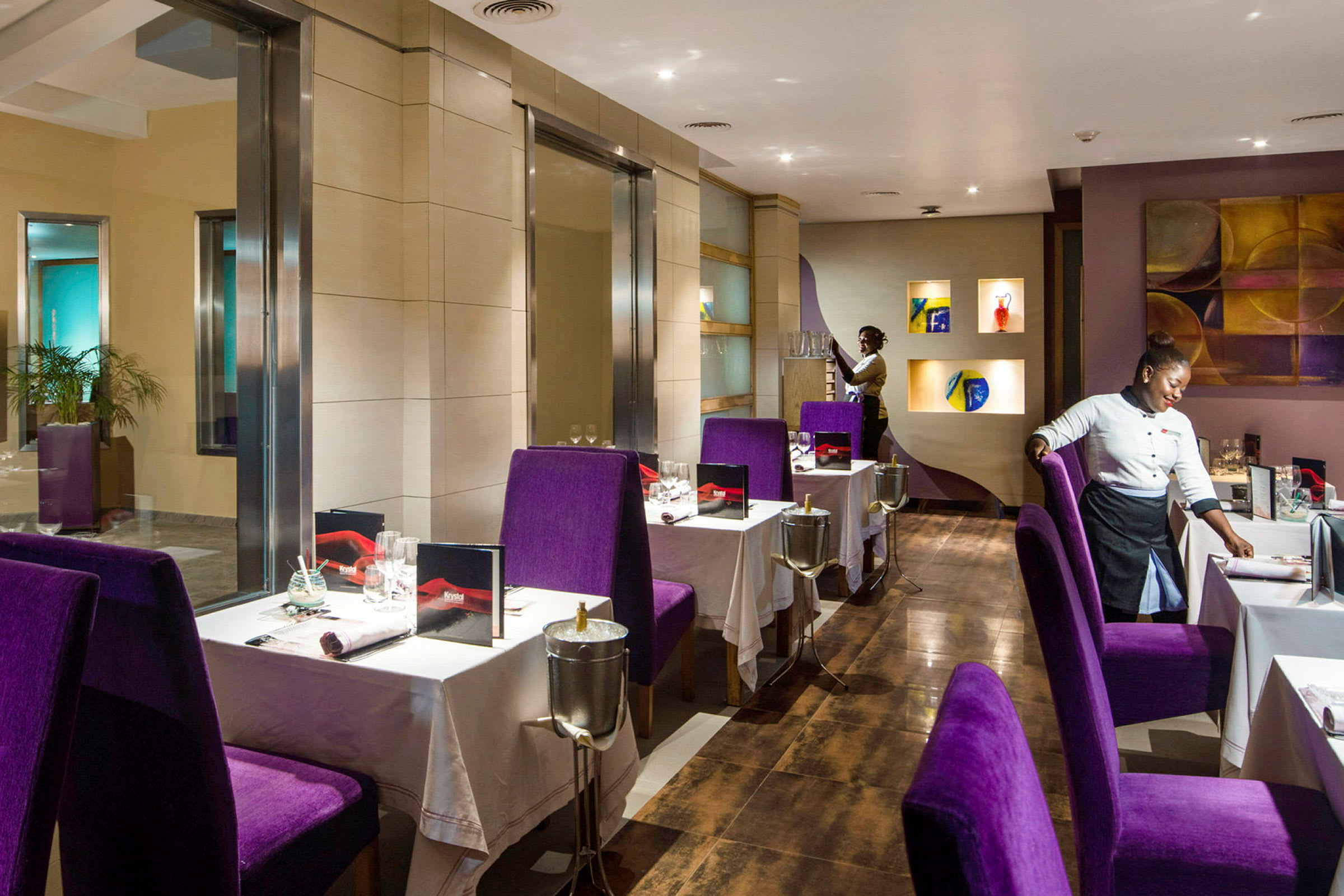 Bild av en elegant inredd restaurang med lila stolar och vita bordsdukar, samt servitörer som förbereder borden. (AI-genererad text)