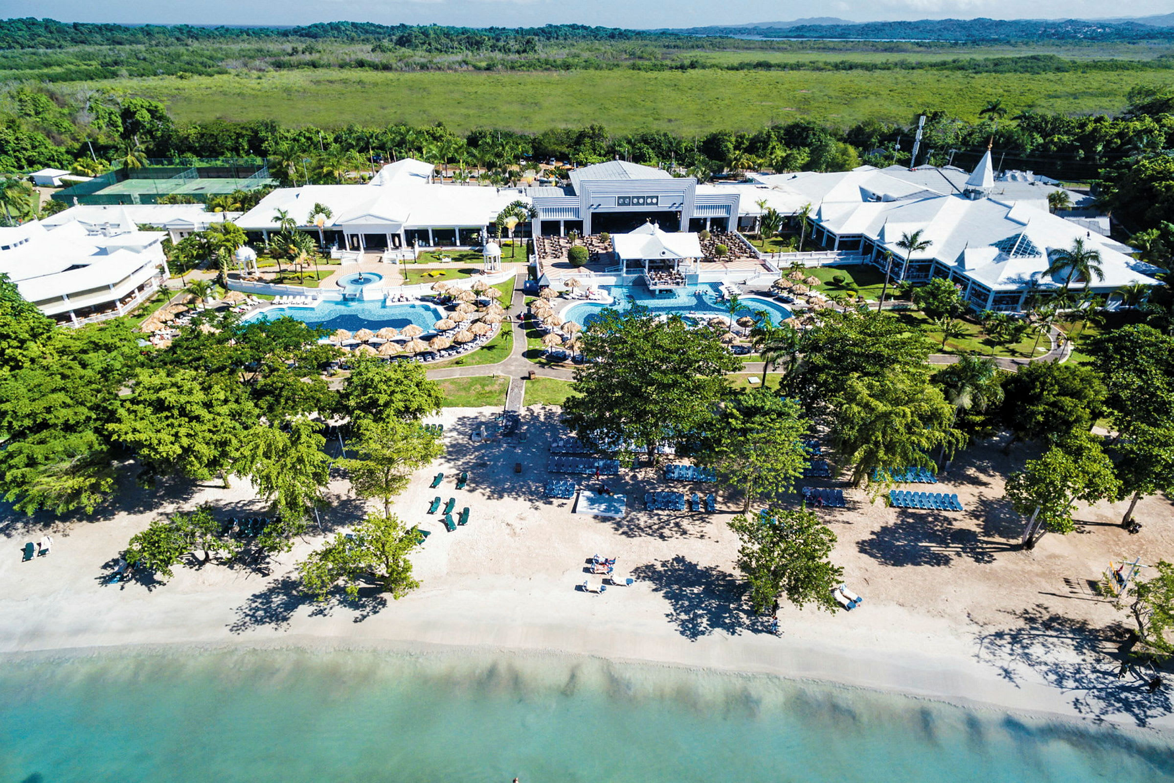 Flygbild av en resort vid stranden med pooler och vit byggnader. (AI-genererad text)