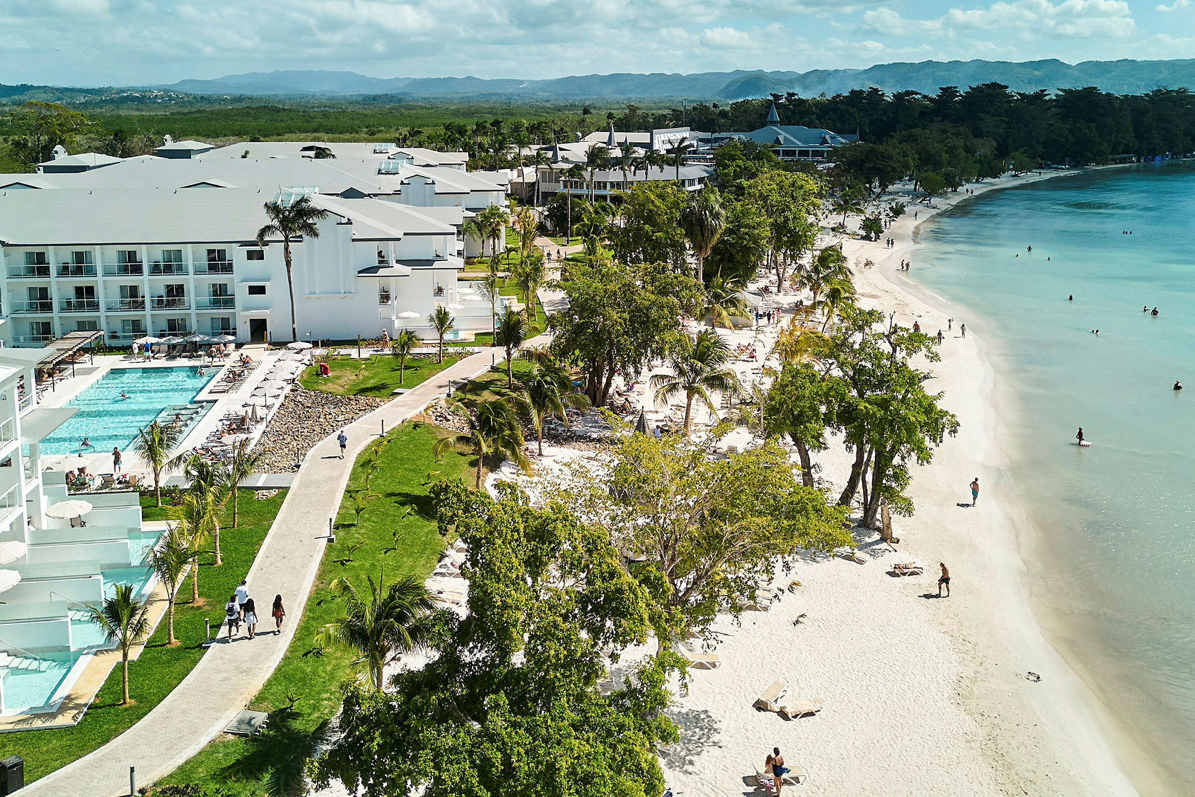 En sandstrand med personer som badar i havet och hotellbyggnader bredvid en pool. (AI-genererad text)