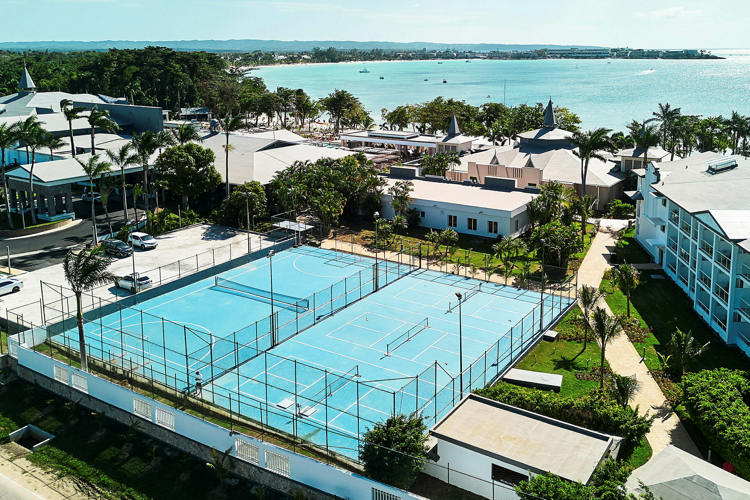 Vy av en tennisbana med blått underlag omgiven av byggnader och grönska, nära havet. (AI-genererad text)