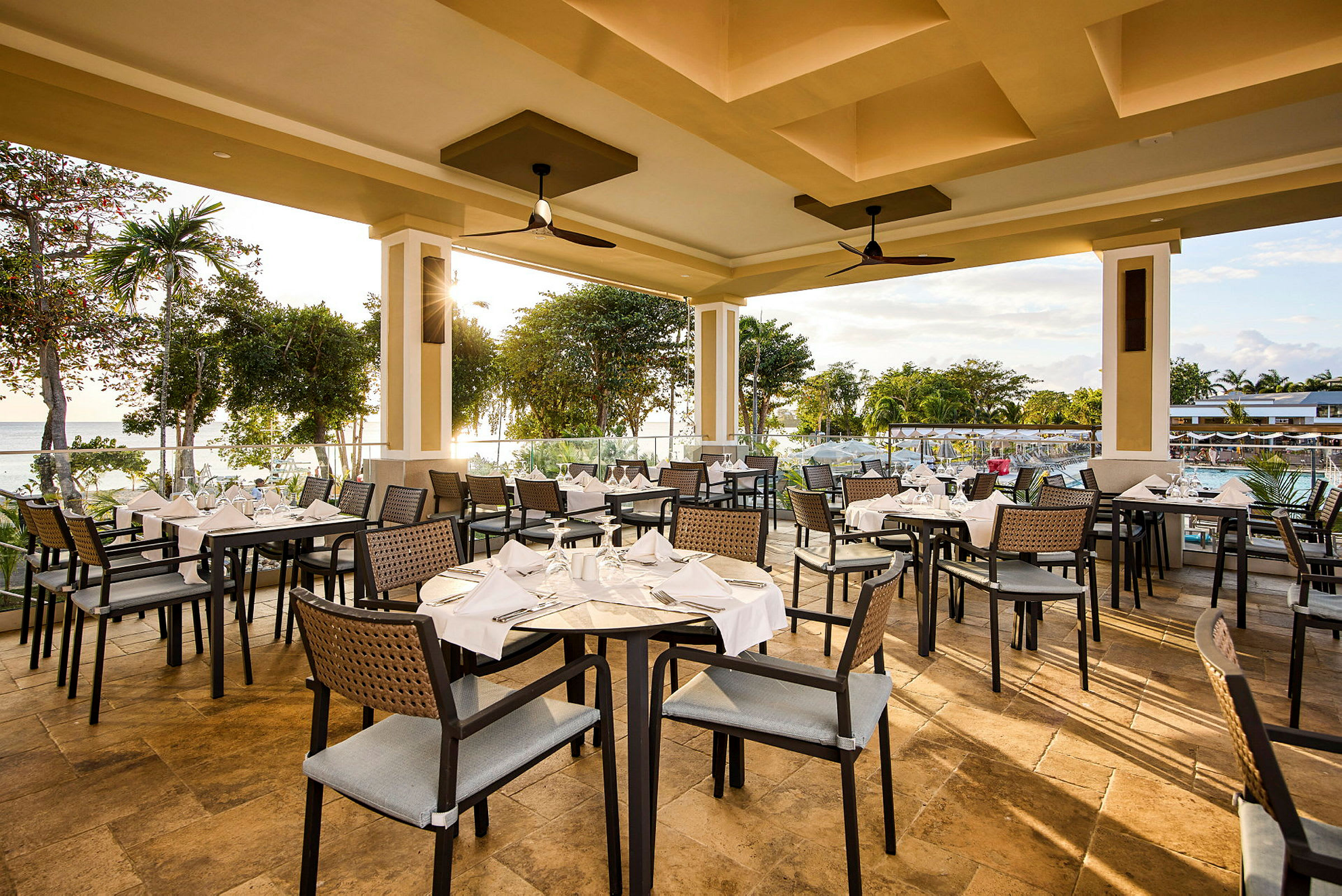 En restaurang med dukade bord och stolar, utsikt över grönska och en swimmingpool. (AI-genererad text)