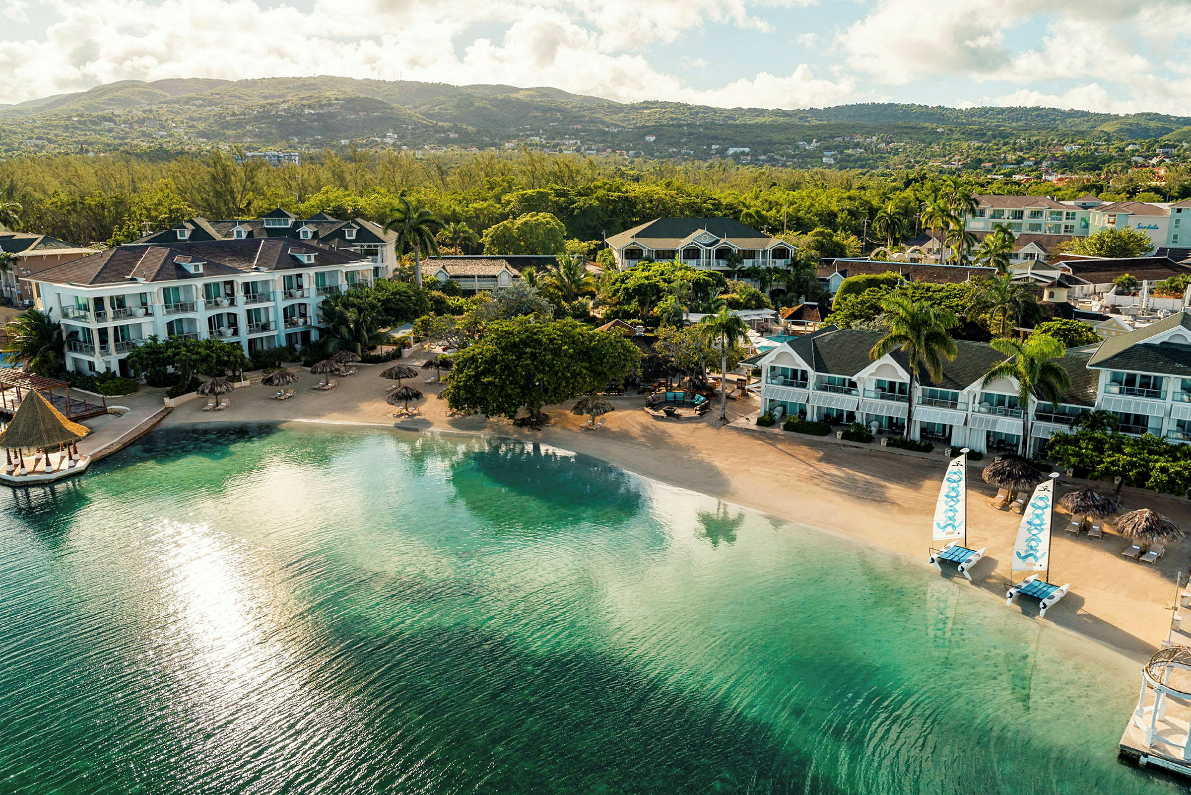 Flera hotellbyggnader vid en lugn sandstrand med klart vatten och utsikt över grönskande kullar. (AI-genererad text)