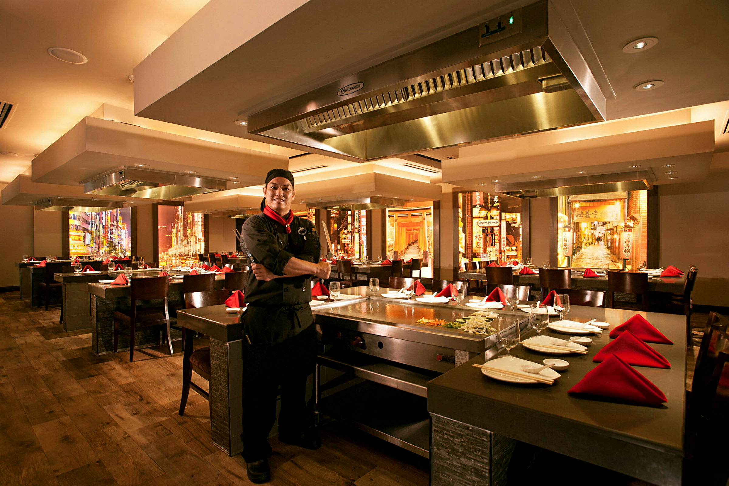 Interiör av en restaurang med uppställda bord och stolar. Ett teppanyaki-kök är synligt med köksutrustning och ingredienser. (AI-genererad text)