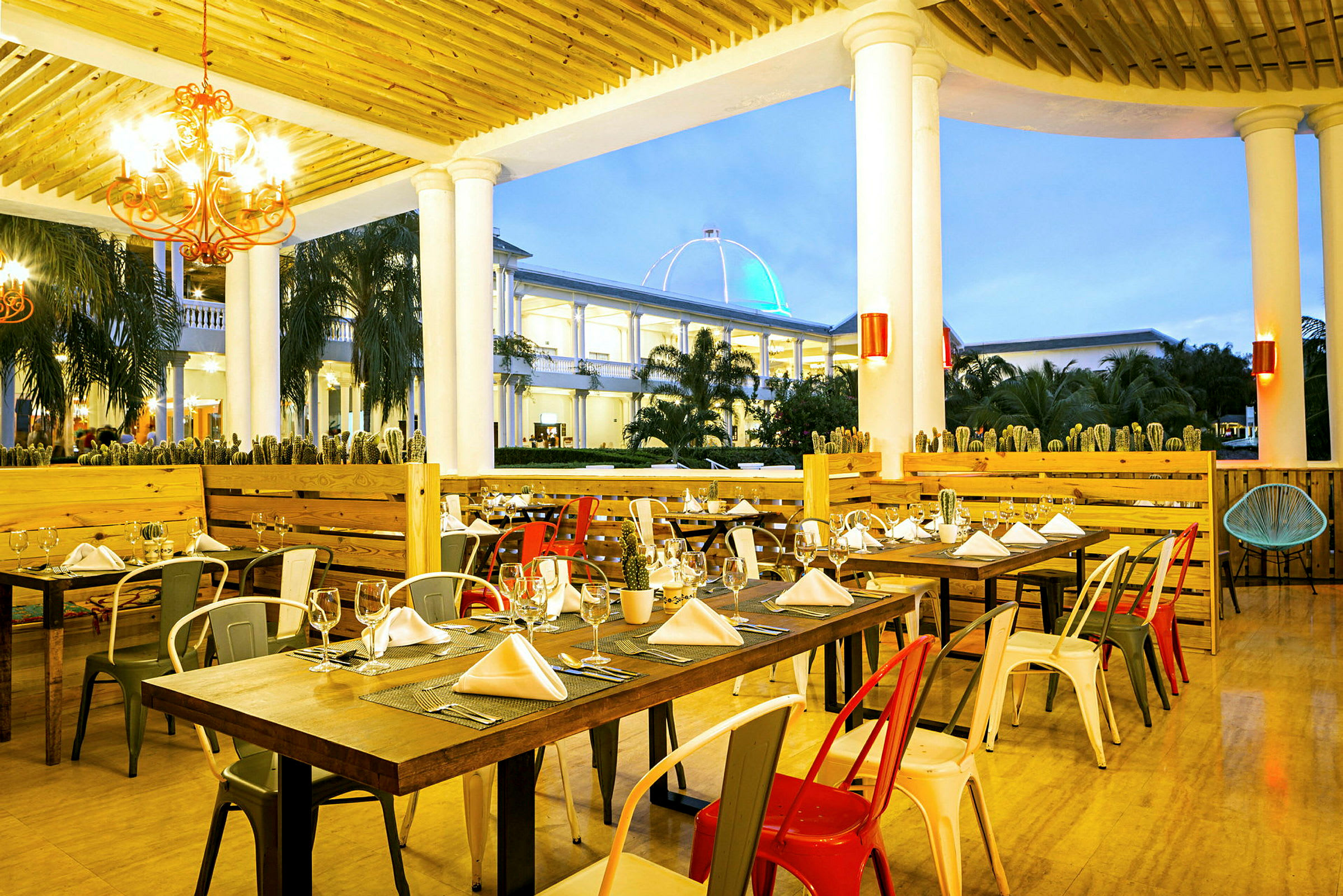 Bild av en restaurang med dukade bord och färgglada stolar, belägen under en träpergola med pelare. I bakgrunden syns en byggnad med ett upplyst kupoltak. (AI-genererad text)