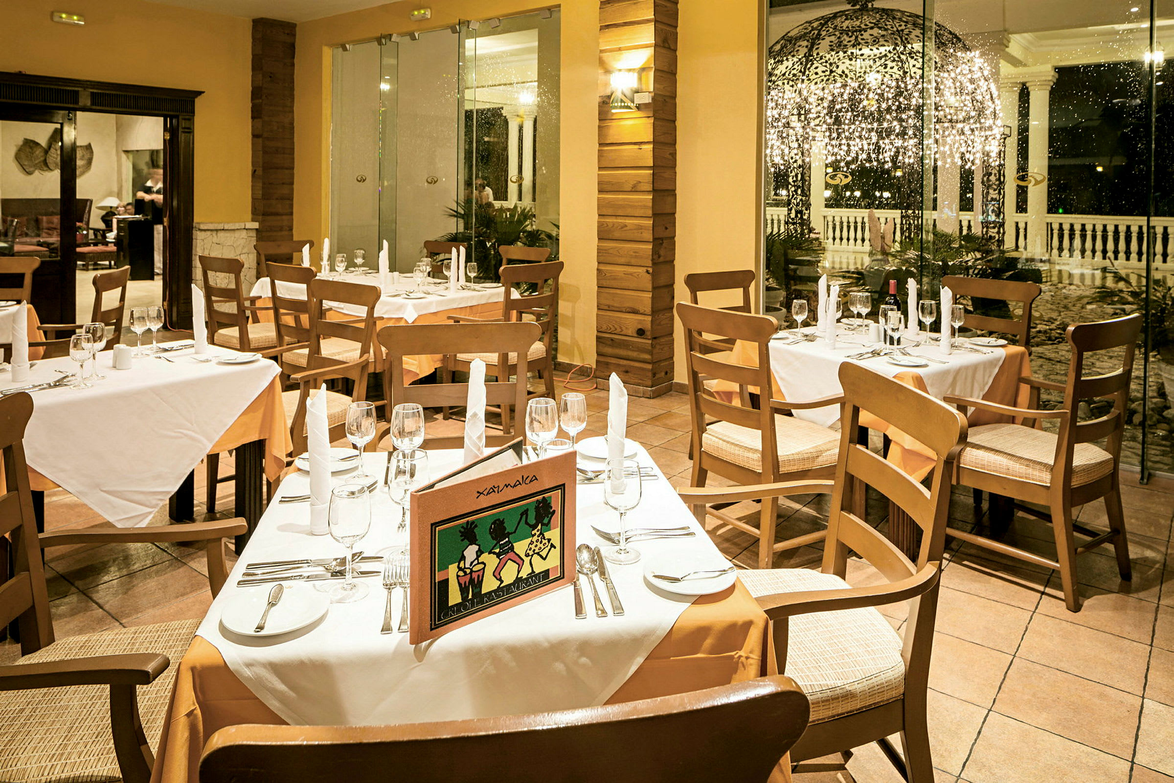 En restaurang med dukade bord, vita dukar och trästolar, med ett öppet område och dekorerat utomhusområde i bakgrunden. (AI-genererad text)