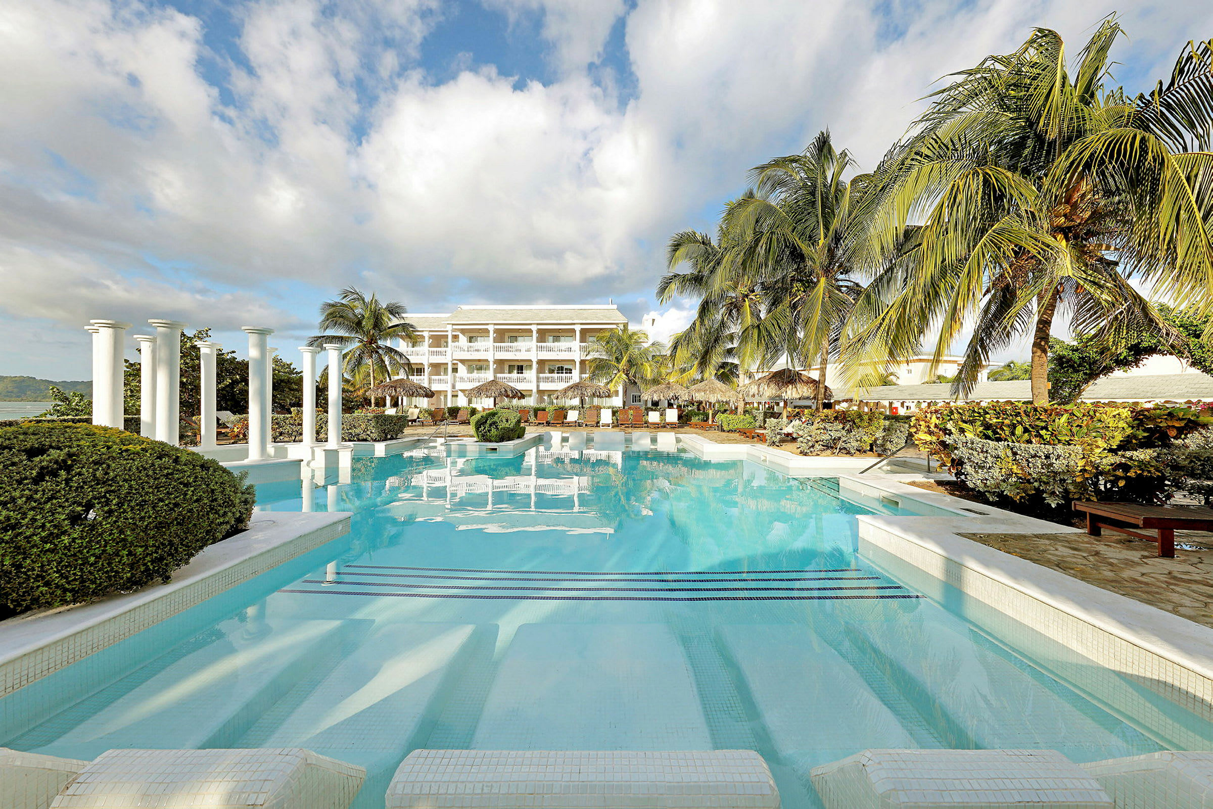 Bild av en pool med omgivande grönska och palmer, samt ett hotellkomplex i bakgrunden. (AI-genererad text)