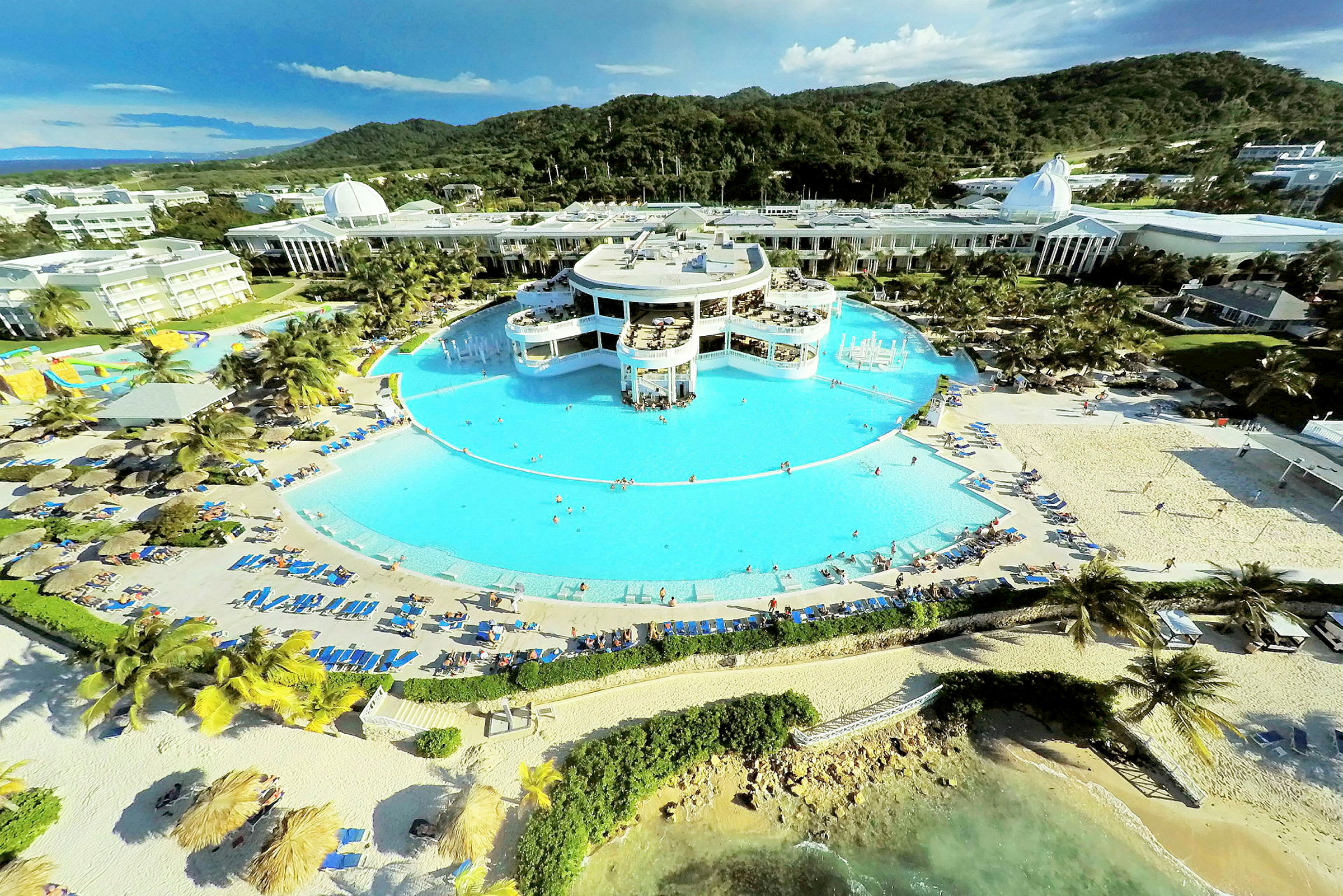 Ett semesterkomplex med en stor rund pool, omgiven av solstolar och sandstrand, med gröna berg i bakgrunden. (AI-genererad text)