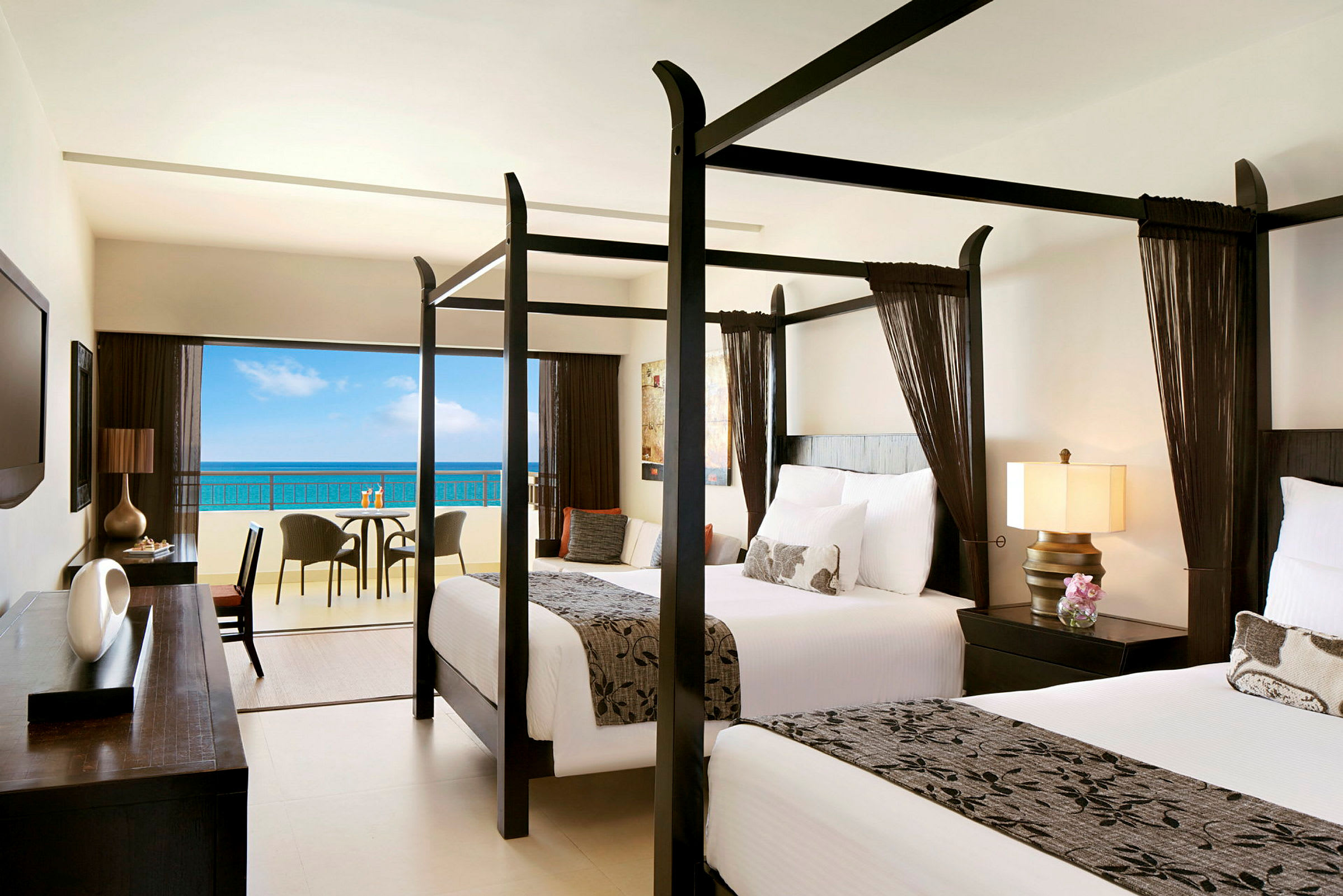 Hotellrum med två sängar med himmelssängar och utsikt över havet från en balkong. (AI-genererad text)