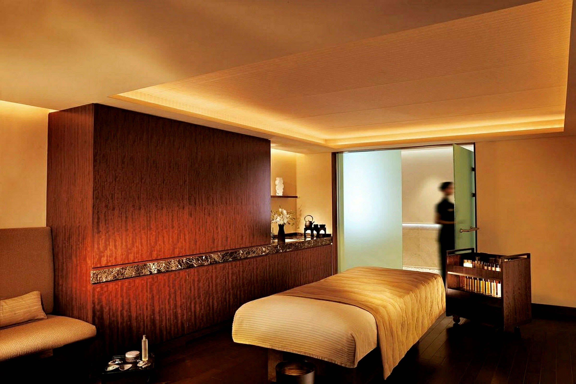 Ett spa-rum med en massagebänk klädd med beige tyg och en mild belysning. Det finns ett sidebord med produkter och en person syns i bakgrunden. (AI-genererad text)