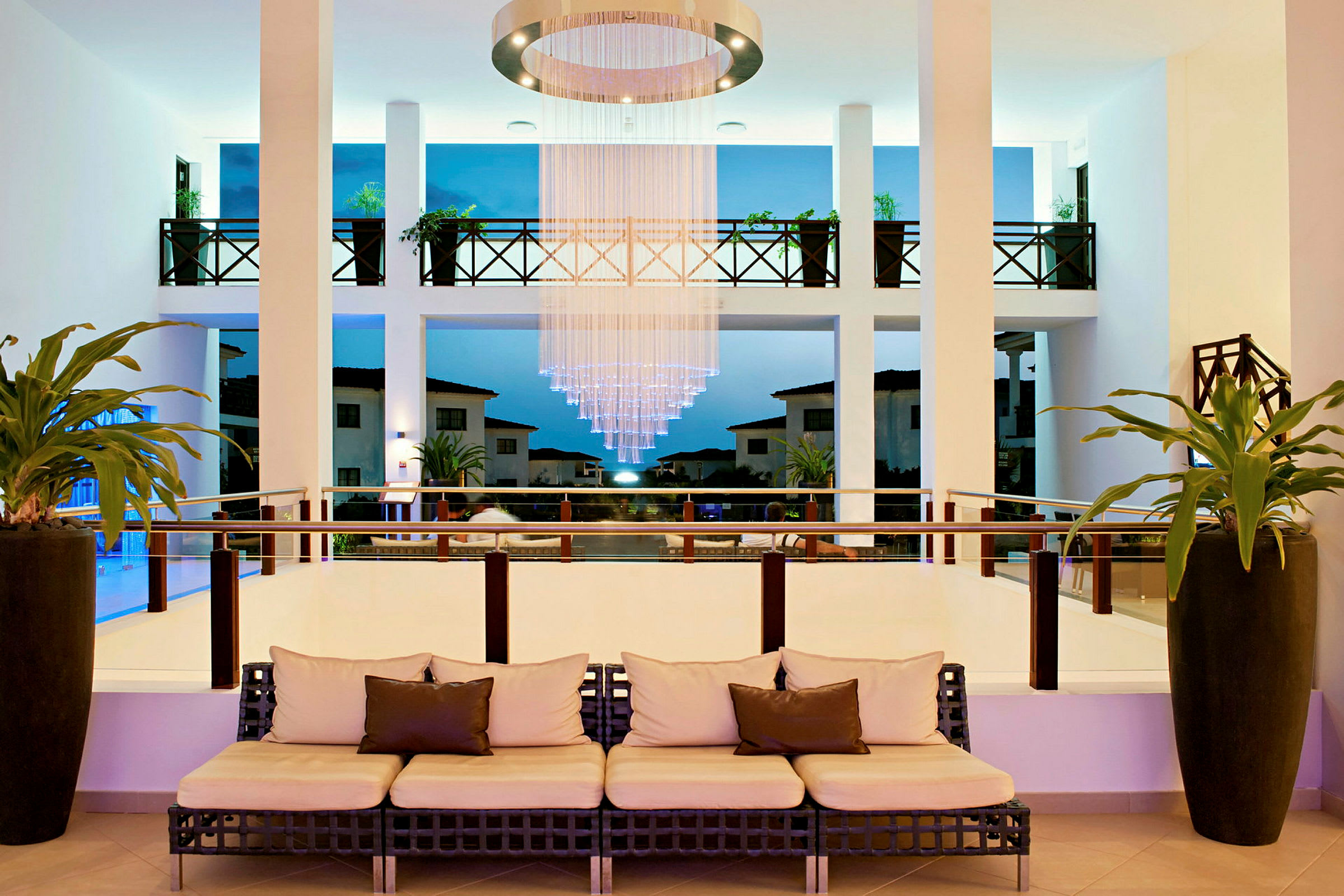 En hotellobby med soffa, hängande ljuskrona och utsikt mot utomhusområde. (AI-genererad text)