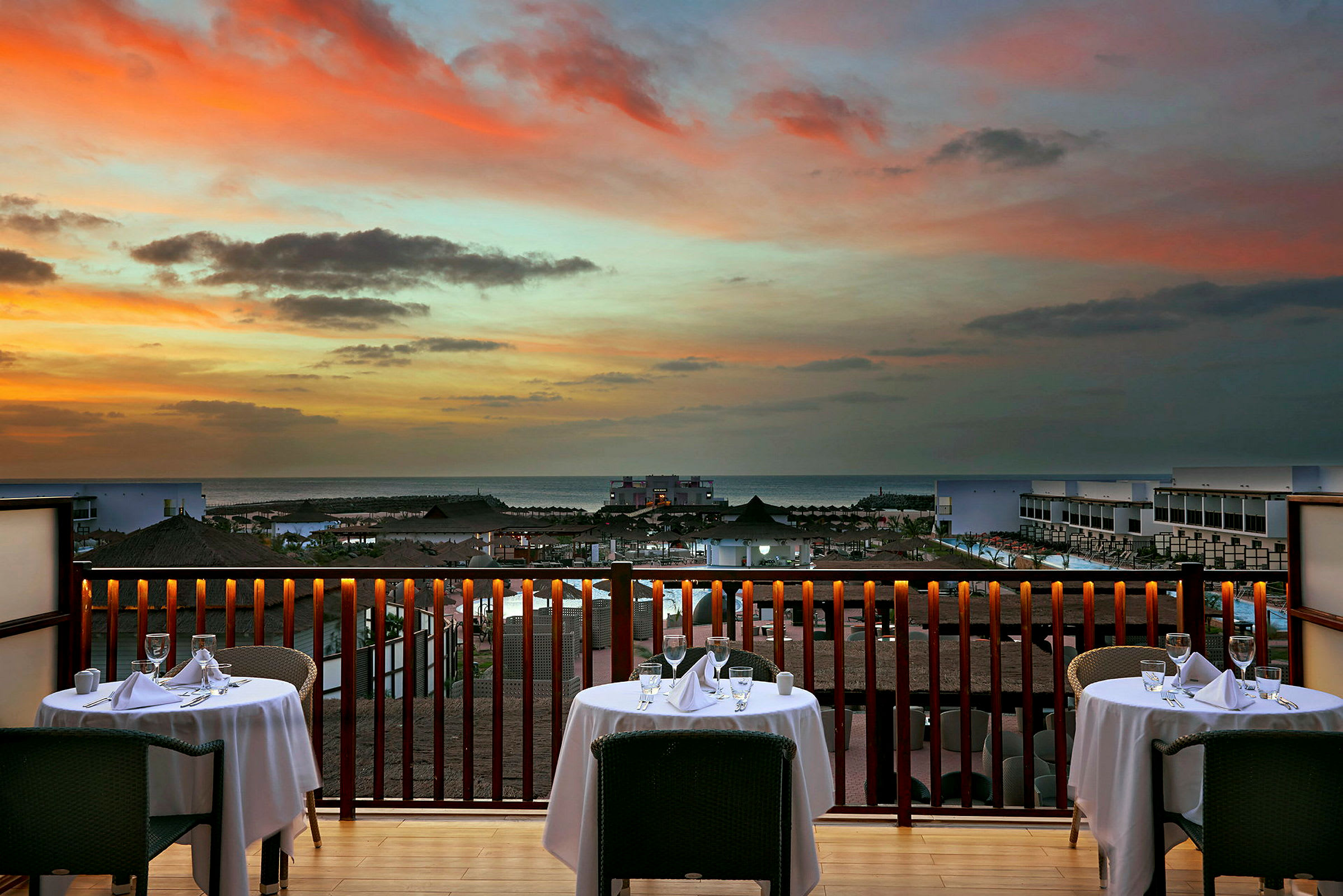 Utsikt från en restaurang med dukade bord mot solnedgången över havet. (AI-genererad text)