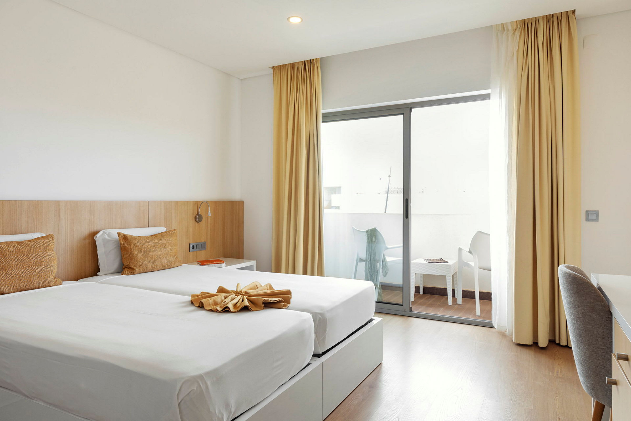 Ett hotellrum med två enkelsängar, guldgula kuddar och en balkong med stolar. (AI-genererad text)