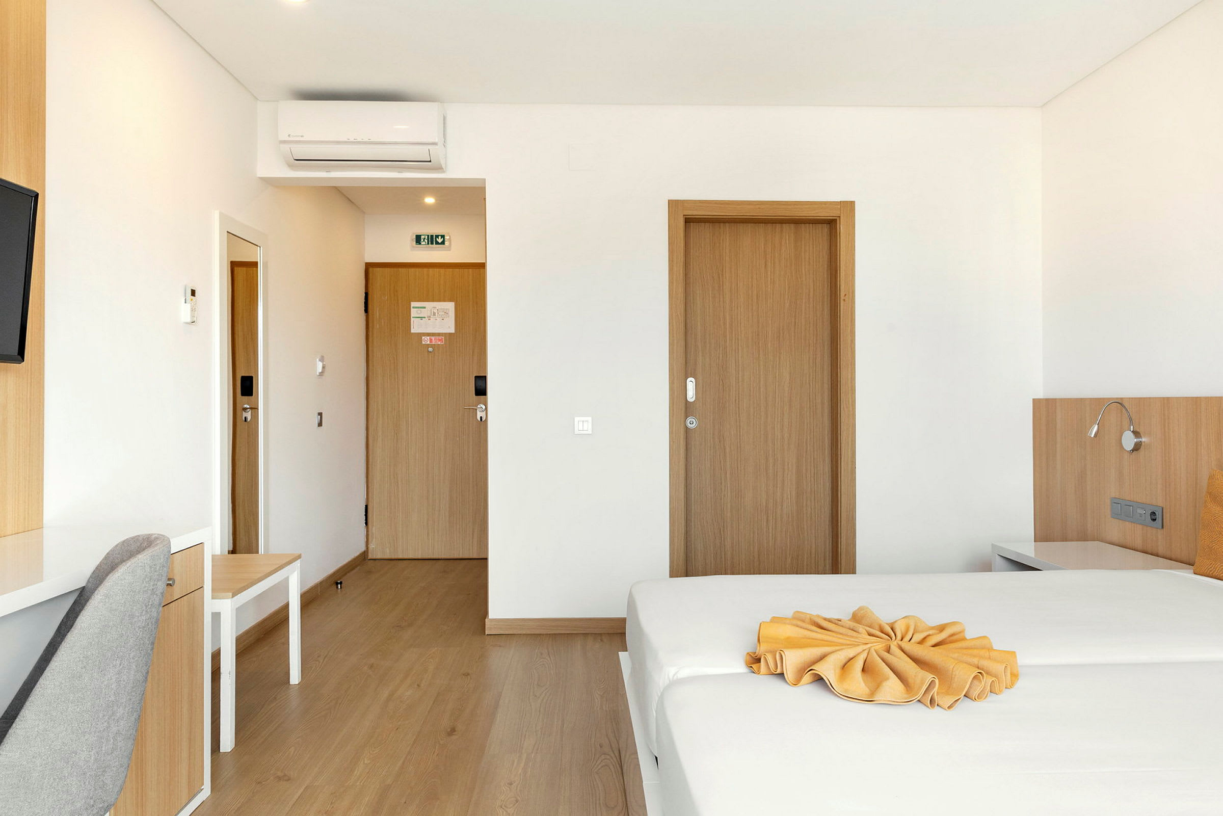 En hotellrum med en bäddad säng och ett skrivbord bredvid en grå stol, dörrar i trä och en luftkonditionering på väggen. (AI-genererad text)