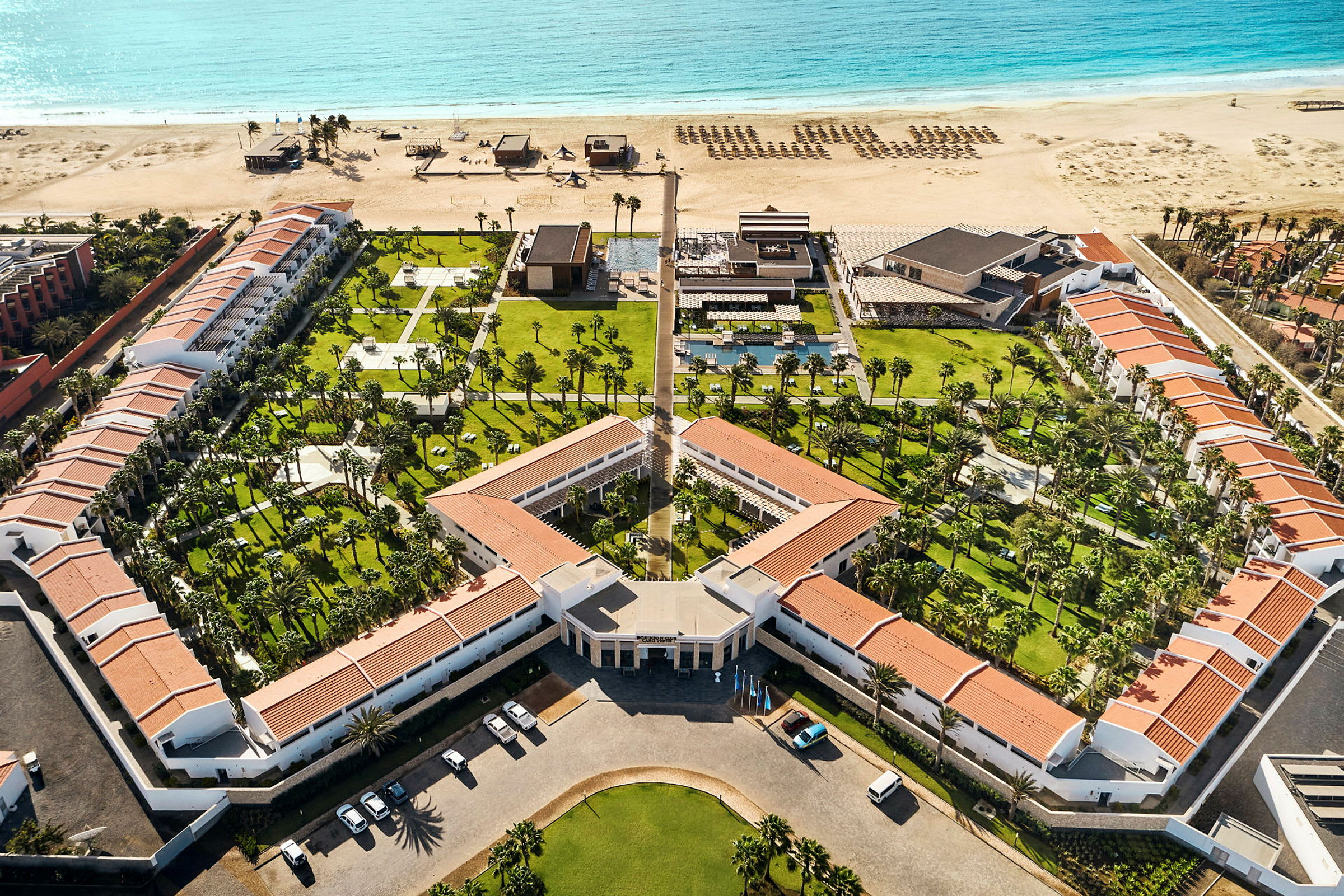 Flygfoto av en strandnära resorts anläggning med poolområden och gröna trädgårdar. (AI-genererad text)
