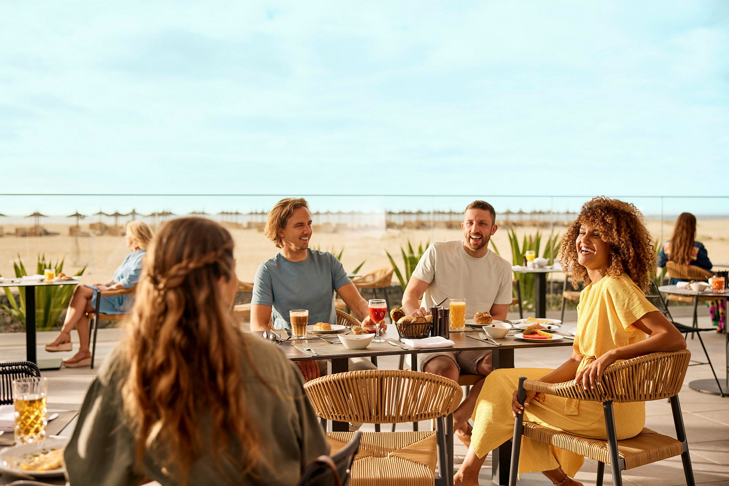 Människor sitter vid bord utomhus och äter måltider, med en strand och havet i bakgrunden. (AI-genererad text)