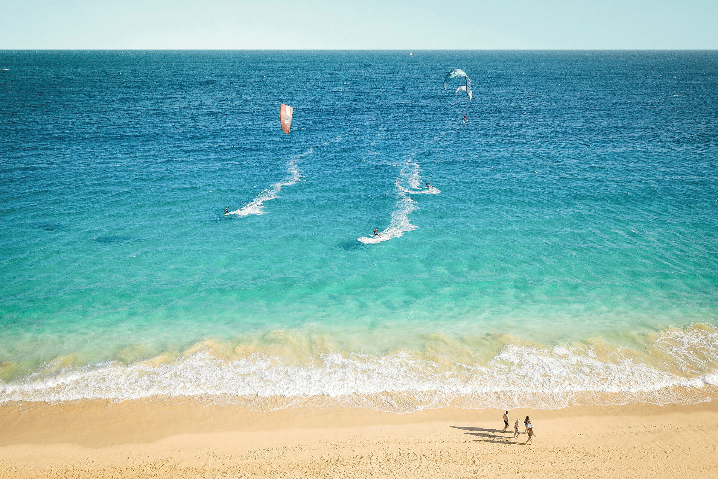 Strand med blått hav där personer kitesurfar och promenerar. (AI-genererad text)