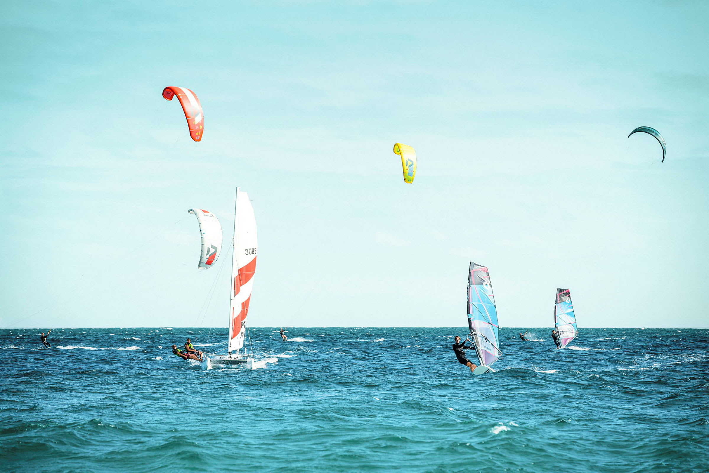 Flera personer deltar i olika vindsportaktiviteter på havet, med segelbåt, kitesurfing och vindbrädor. (AI-genererad text)