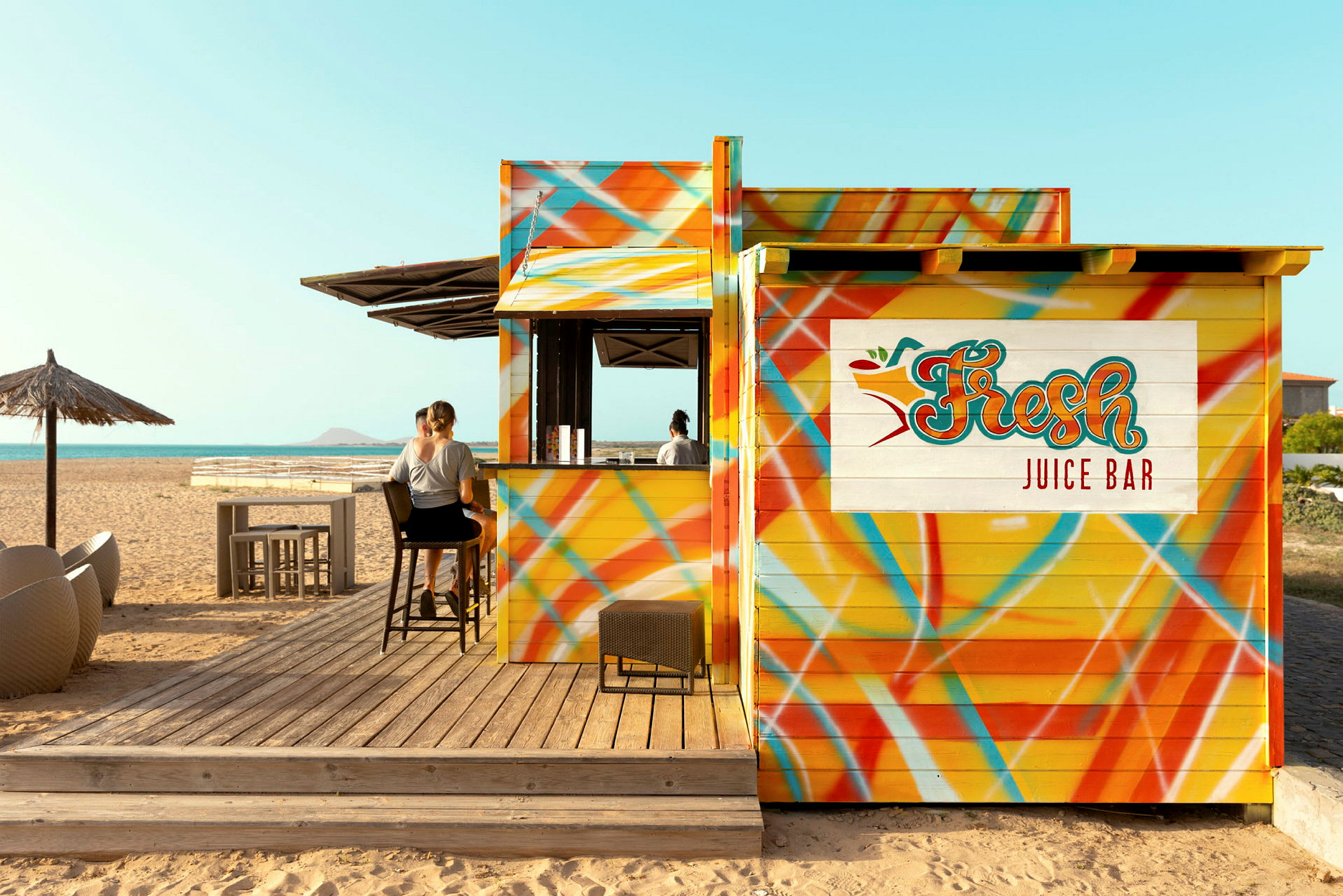 En färgglad juicebar vid stranden med personer som sitter på barstolar. (AI-genererad text)