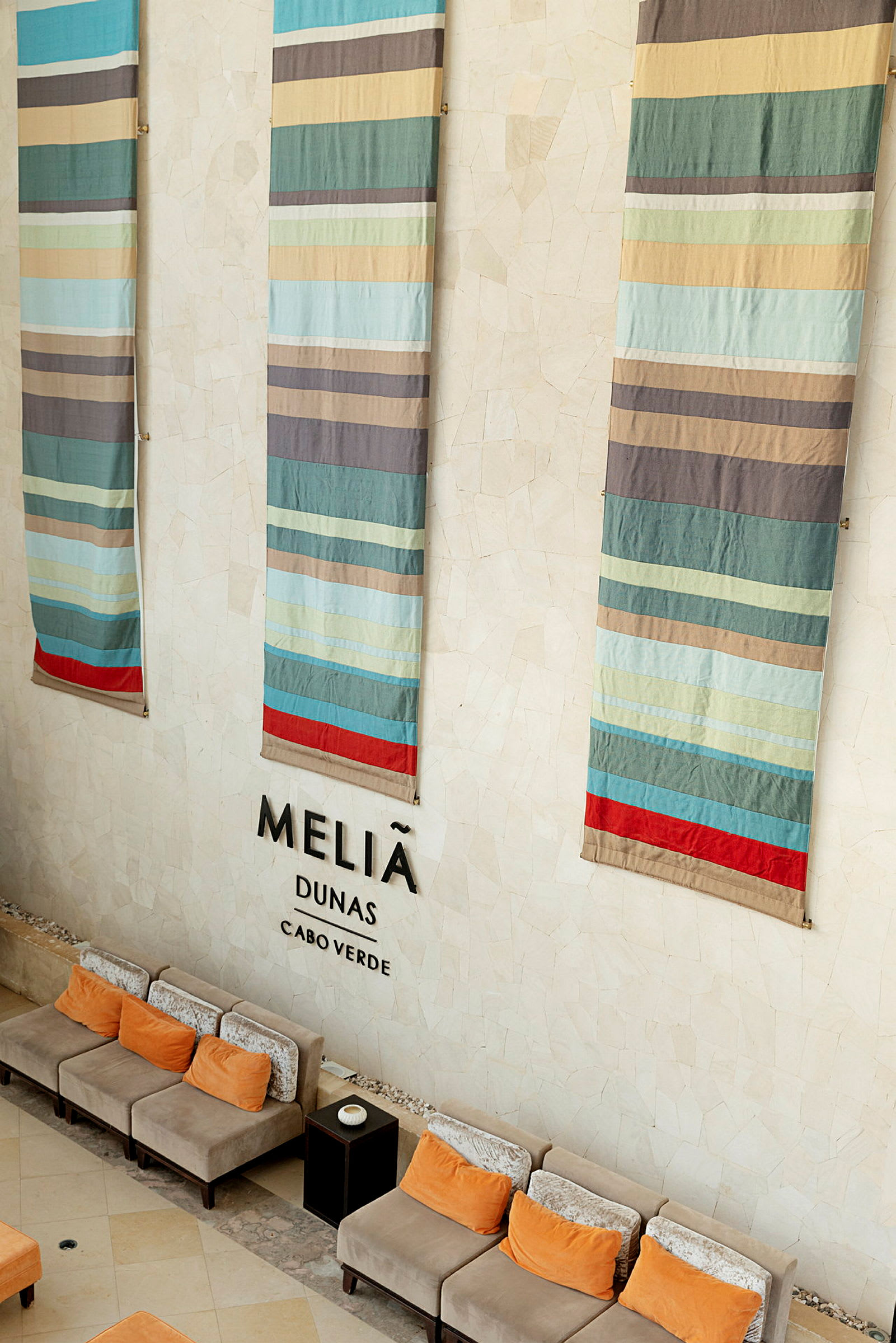 Dekorativa tygstycken hänger på en hotellvägg med texten 'Meliã Dunas Cabo Verde', och sittplatser med orange kuddar syns nedanför. (AI-genererad text)
