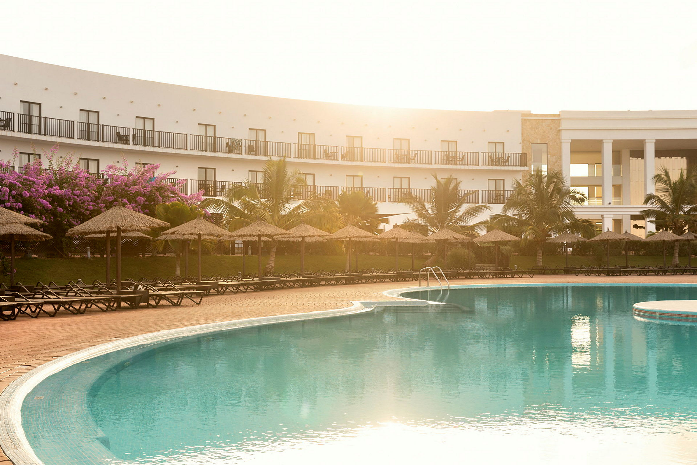 Hotellpoolområde med parasoller och solstolar framför en vit byggnad. (AI-genererad text)