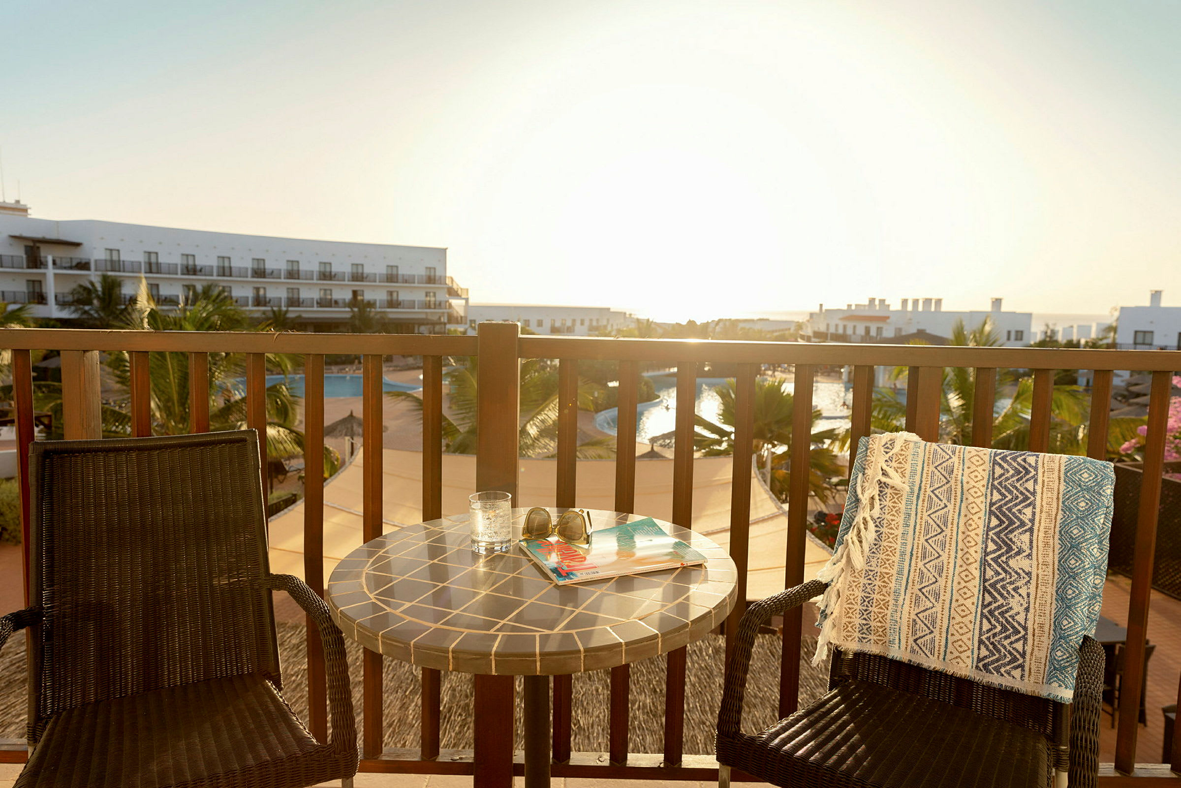 En balkong med två stolar, ett bord och utsikt över en pool och hotellbyggnader. (AI-genererad text)