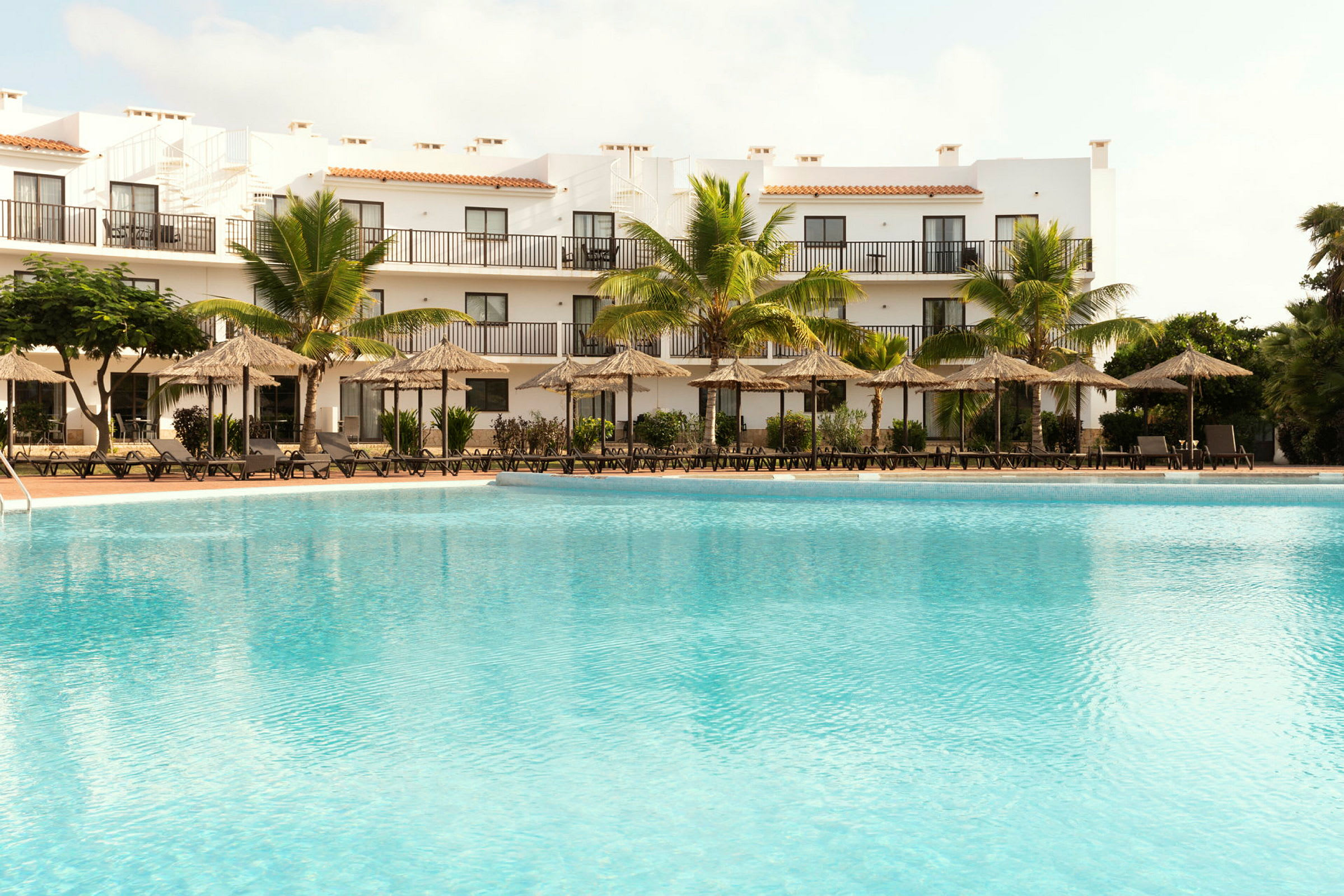 Pool med solstolar framför ett hotell med balkonger och palmträd. (AI-genererad text)