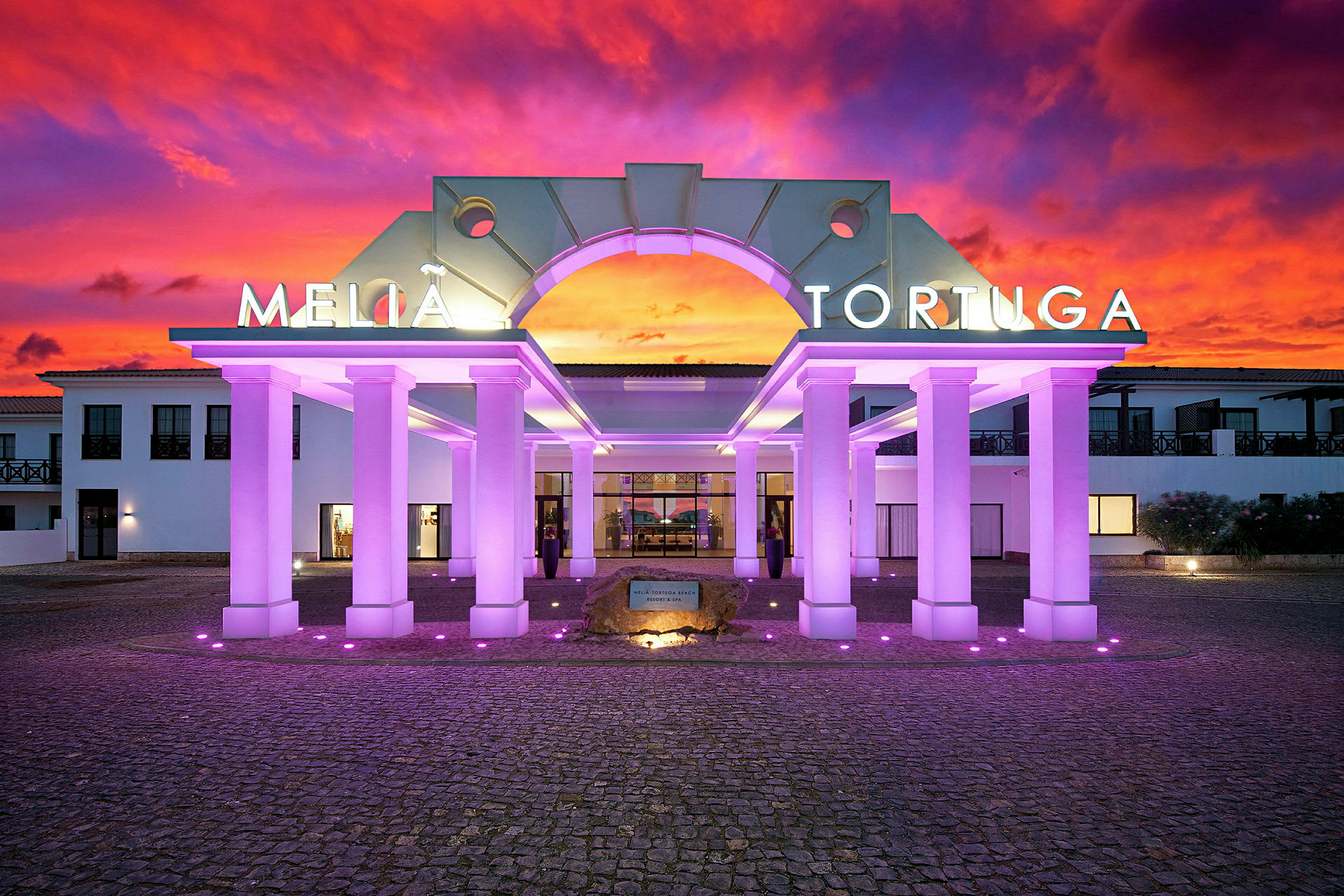 Ingången till hotellet med upplysta pelare och en färgstark solnedgång i bakgrunden. (AI-genererad text)