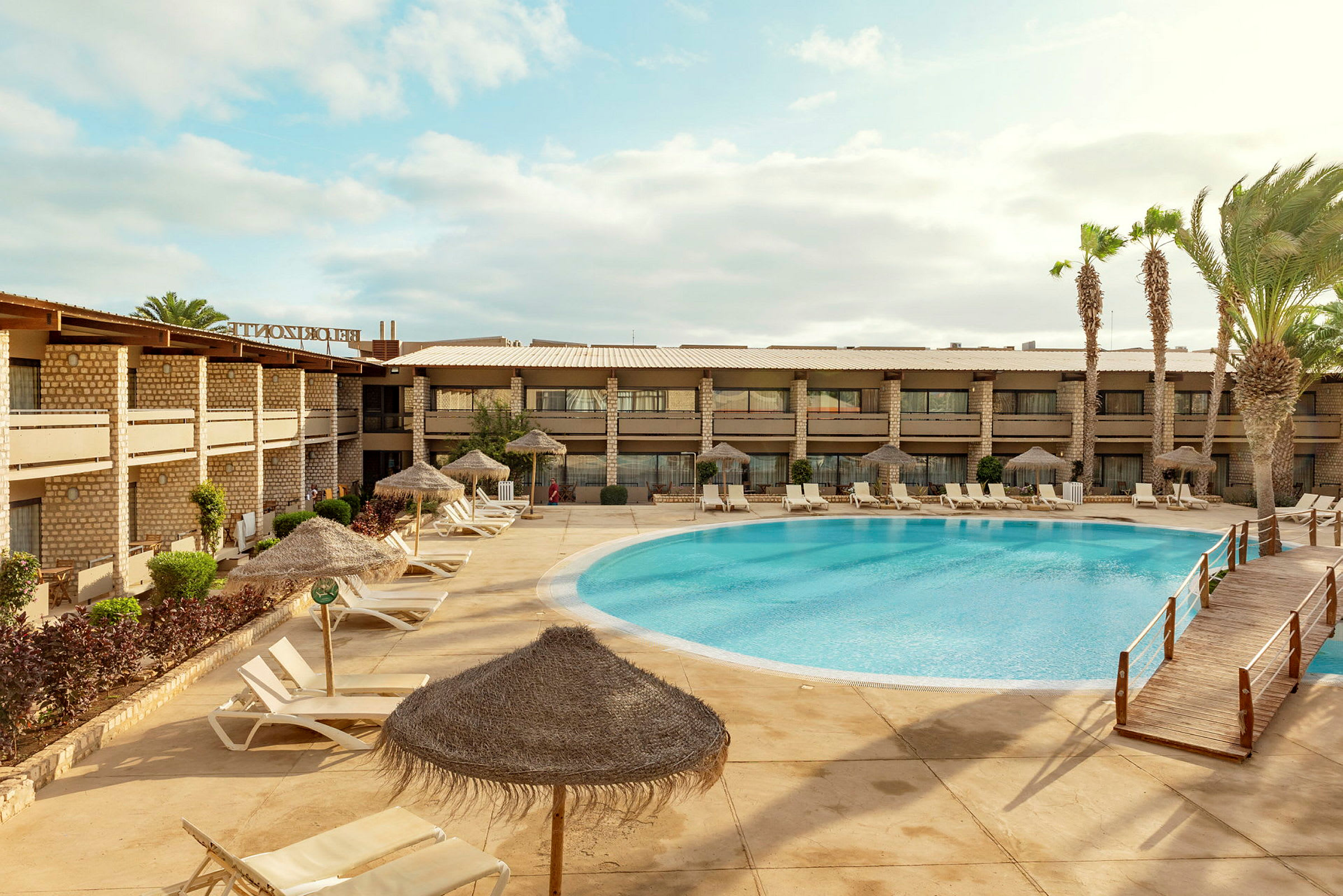 Hotellbyggnad vid en utomhuspool med solstolar och parasoller. (AI-genererad text)