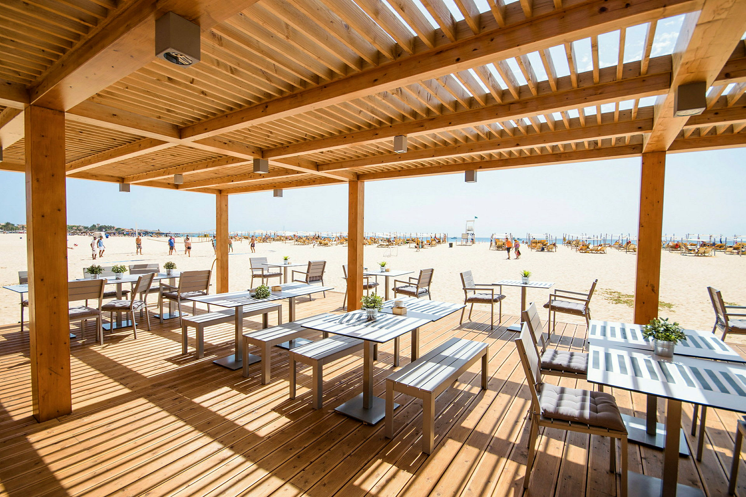Ett träpergola över uteservering med utsikt över en sandstrand. (AI-genererad text)