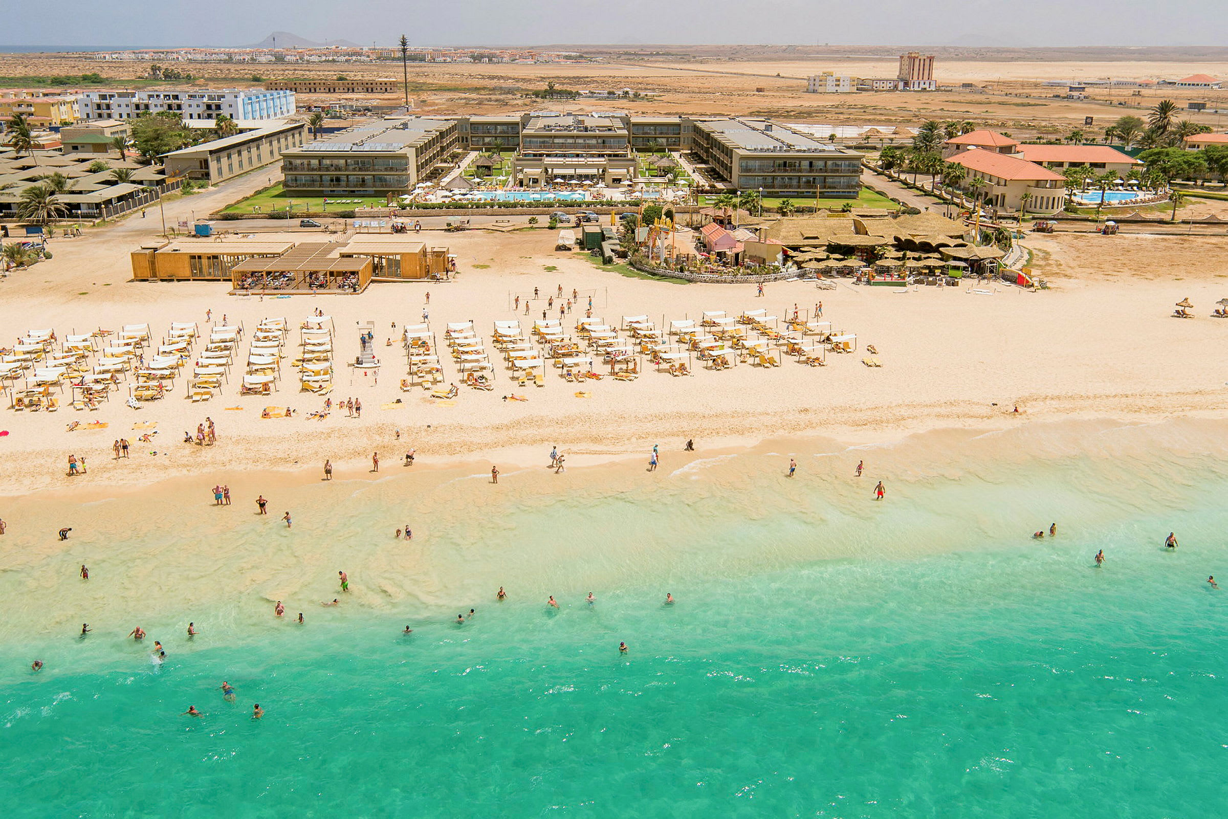 Flygfoto av en strand med turkost vatten, solstolar och parasoller, samt bakom en hotellbyggnad. (AI-genererad text)