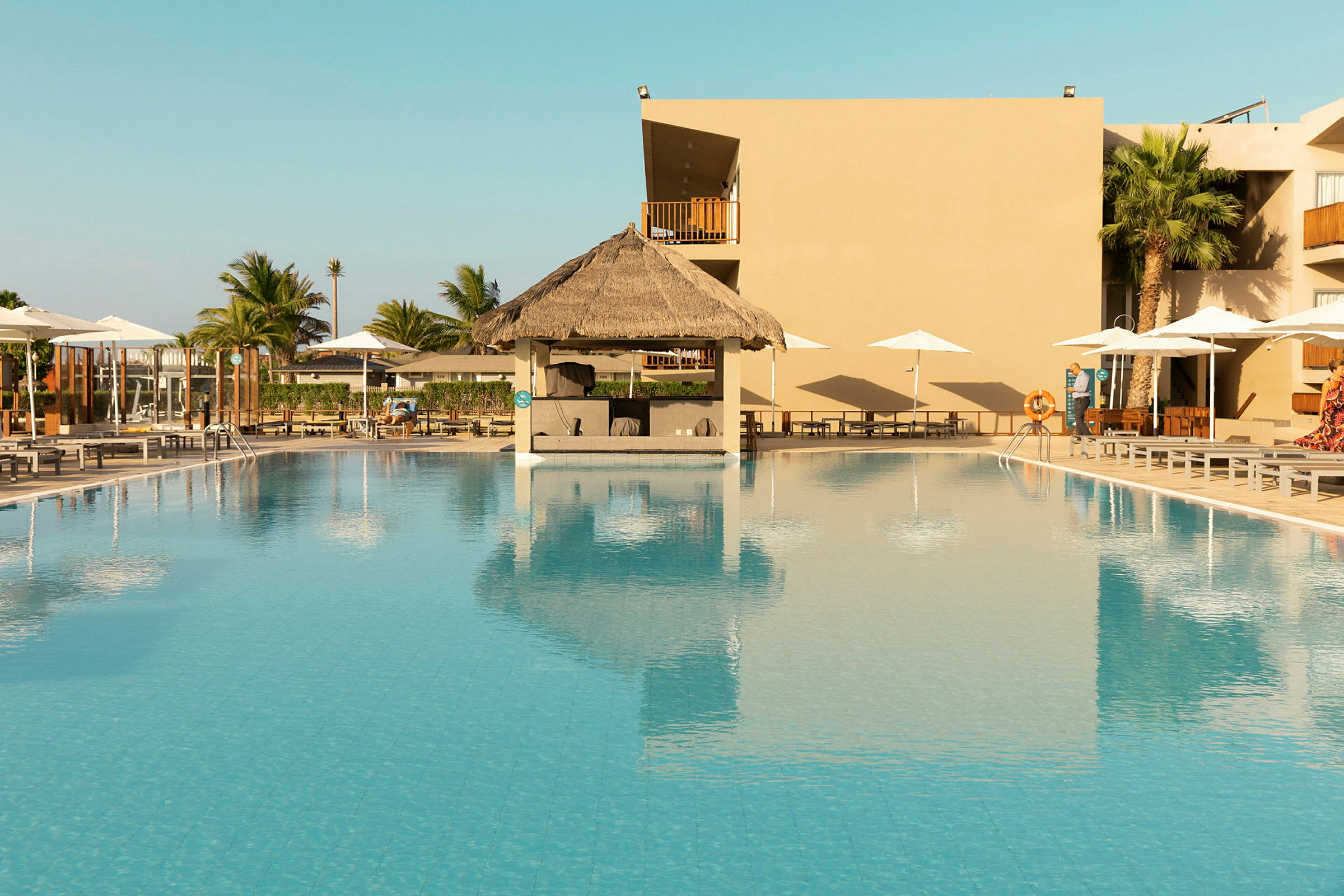 Pool med parasoller och ett mindre taktäckt område med halmtak. (AI-genererad text)