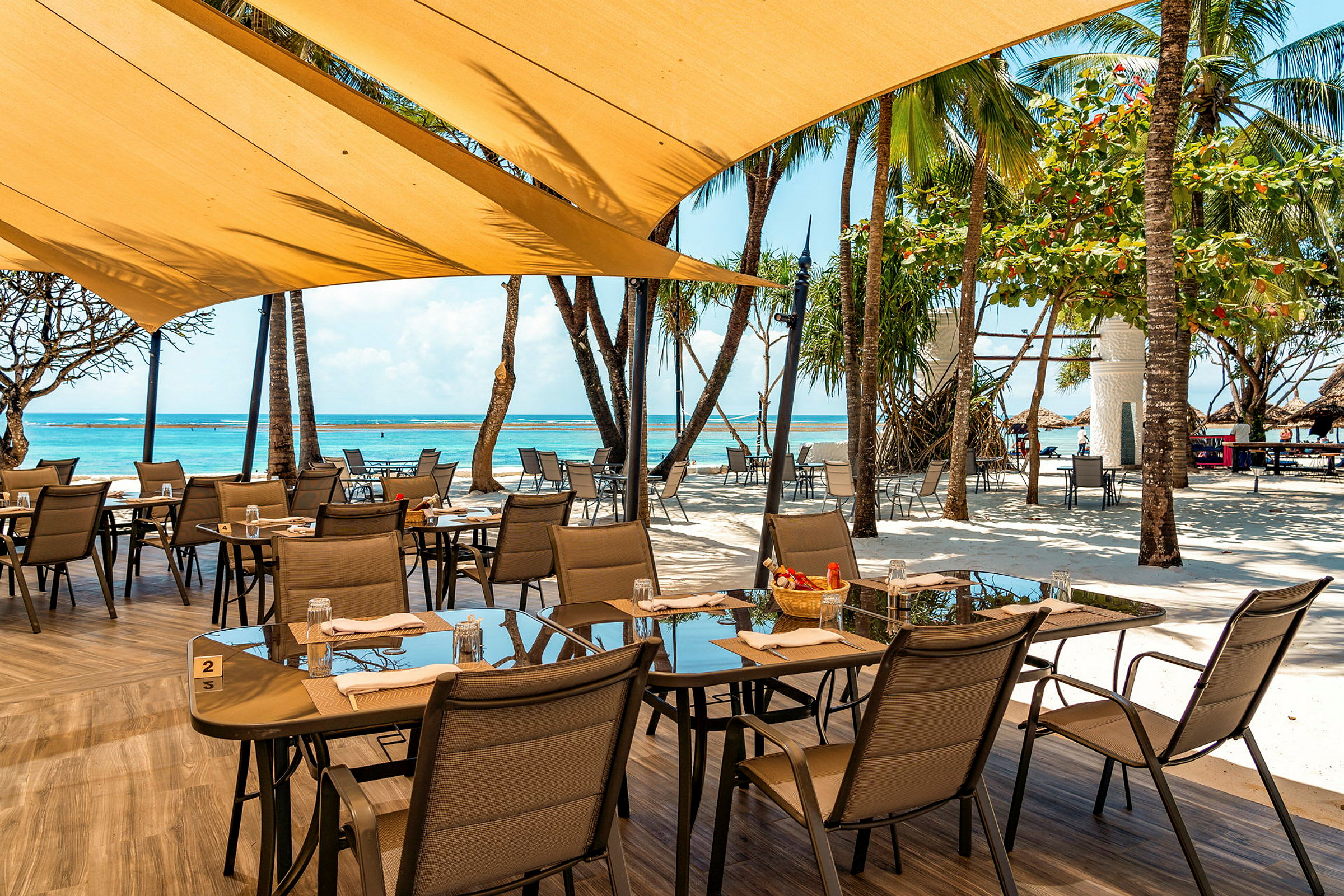 Utomhusrestaurang med dukade bord vid en sandstrand. Möblerna består av glasbord och stolar med solskydd ovanför. (AI-genererad text)